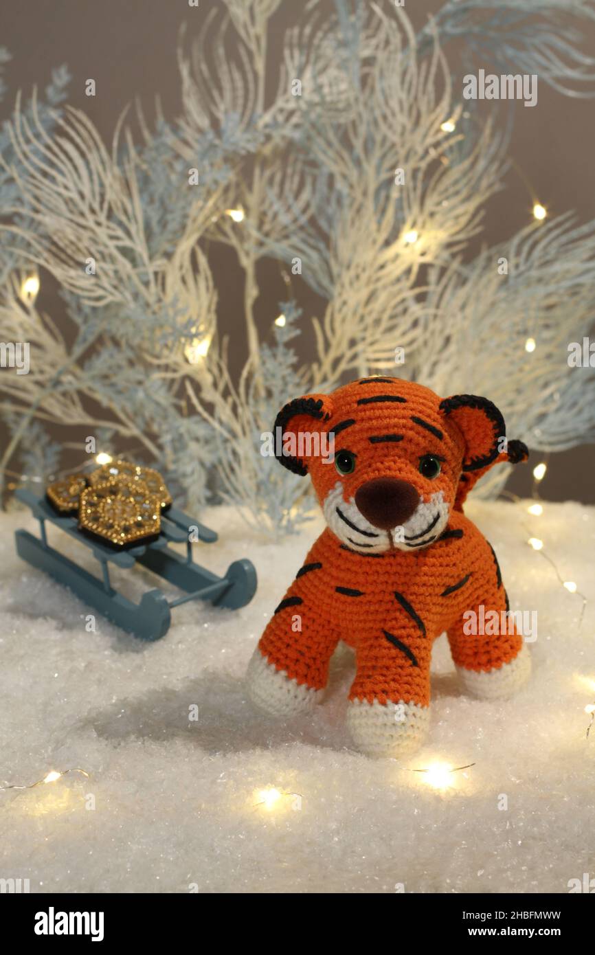 Un cucciolo di tigre di lana lavorato a maglia si trova accanto agli alberi blu chiaro giocattolo con luci e slitte giocattolo con monete su neve bianca artificiale su sfondo grigio. Chiudi Foto Stock