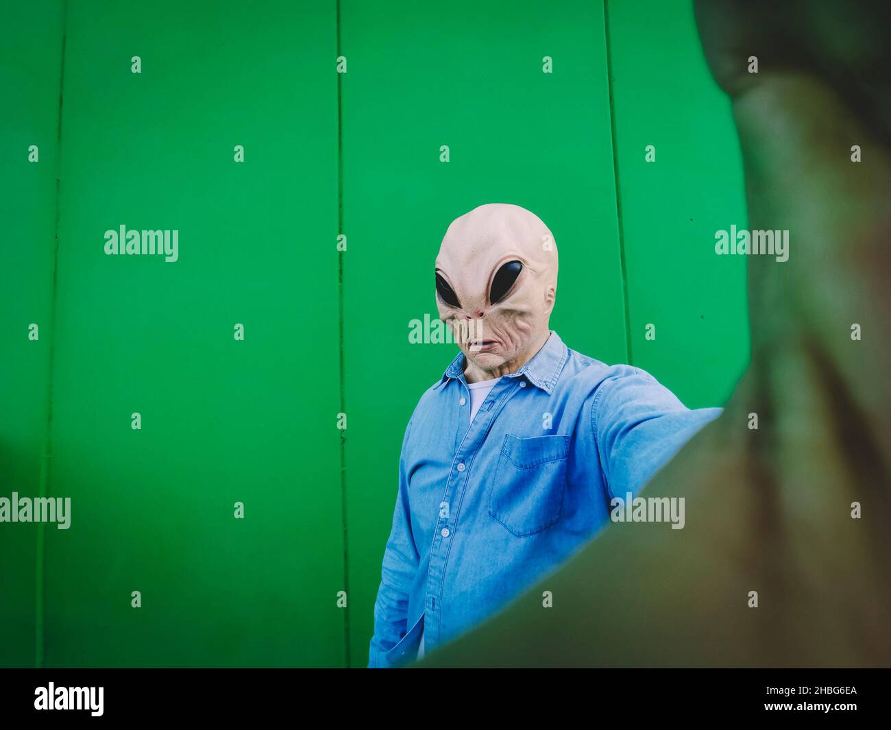 Maschera aliena in piedi faccia scattare foto selfie contro la parete di sfondo verde. Copia spazio per divertente fumetto pubblicità. UFO incasuale extraterrestre de Foto Stock