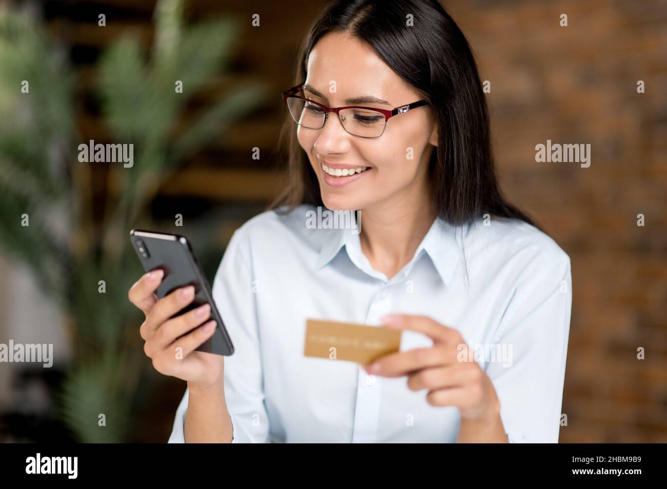 Positivo caucasico giovane adulto bruna donna indossare occhiali, utilizzando smartphone e carta di credito per pagare gli acquisti e la consegna su Internet, inserendo i dati della carta di credito, guardando il telefono, sorridendo Foto Stock