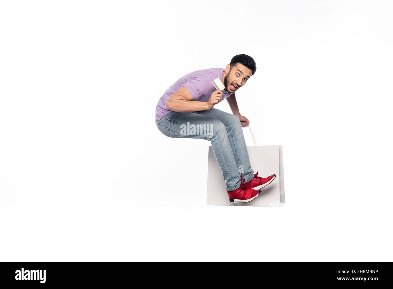 l'uomo stupito in jeans e t-shirt viola levitating mentre tiene la carta di credito e la borsa della spesa su bianco Foto Stock