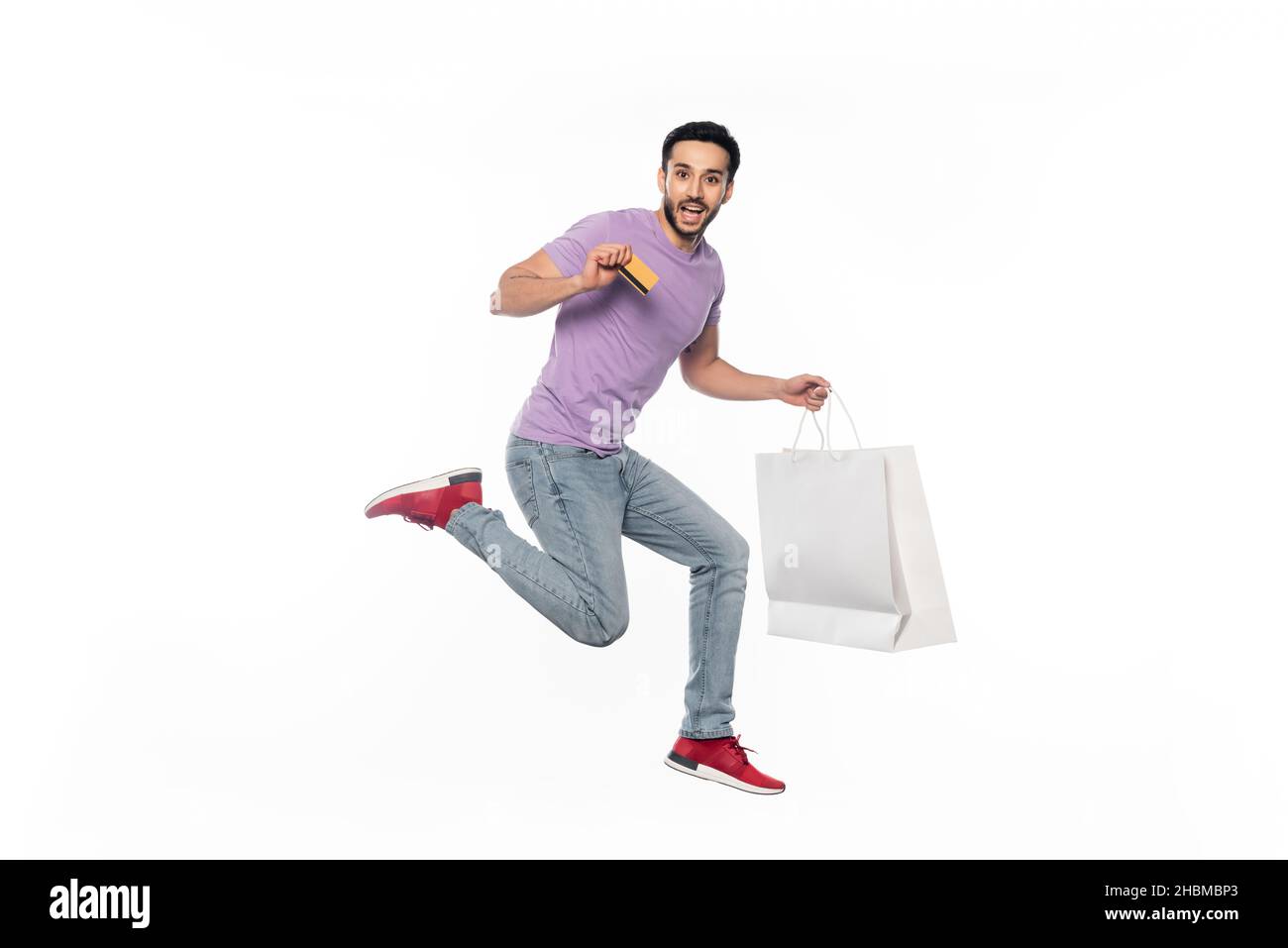 l'uomo eccitato in jeans e t-shirt viola levitating mentre tiene la carta di credito e la borsa della spesa su bianco Foto Stock