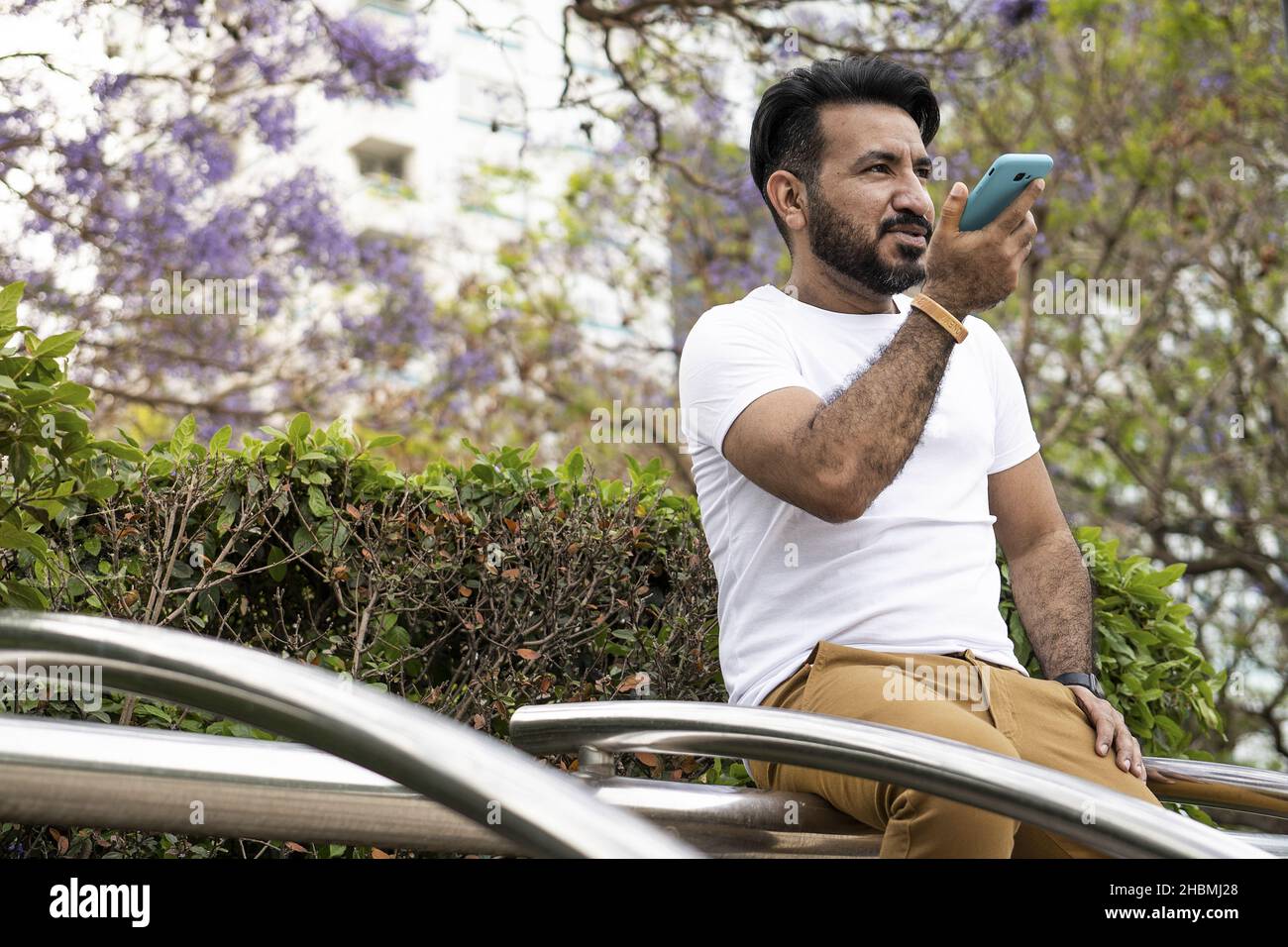 Un uomo ispanico di mezza età seduto su una struttura tubolare in acciaio e che registra un messaggio vocale Foto Stock