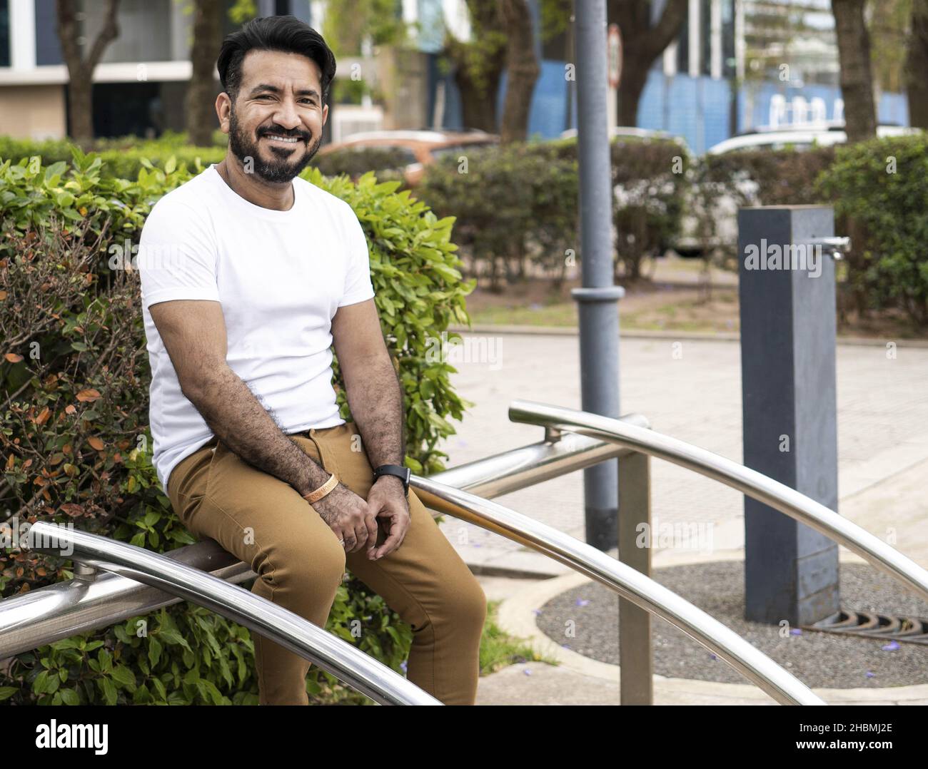 Un uomo ispanico di mezza età seduto su una struttura tubolare in acciaio in un parco e sorridente Foto Stock