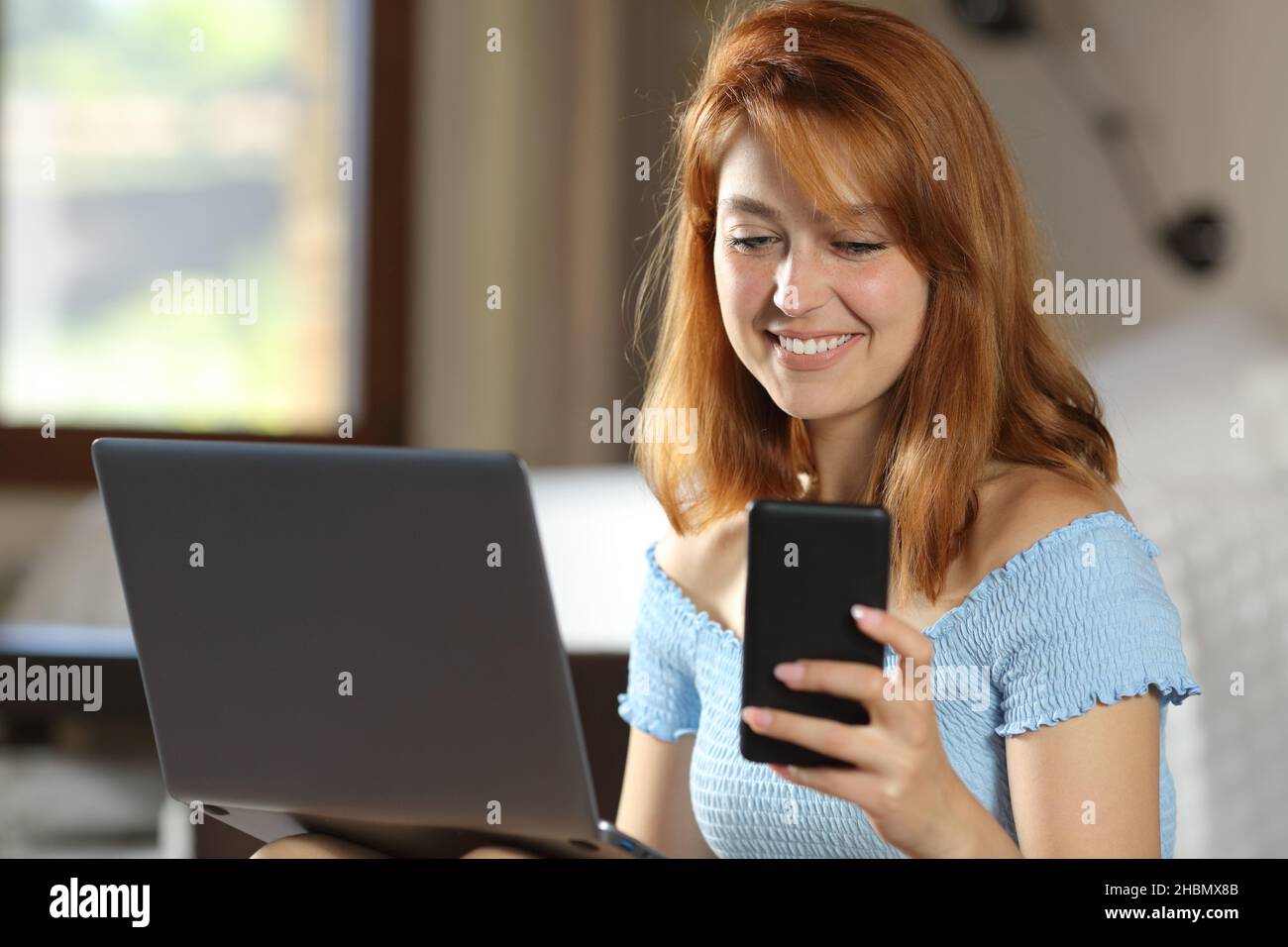 Donna felice che usa il portatile e il telefono cellulare in una stanza della casa Foto Stock