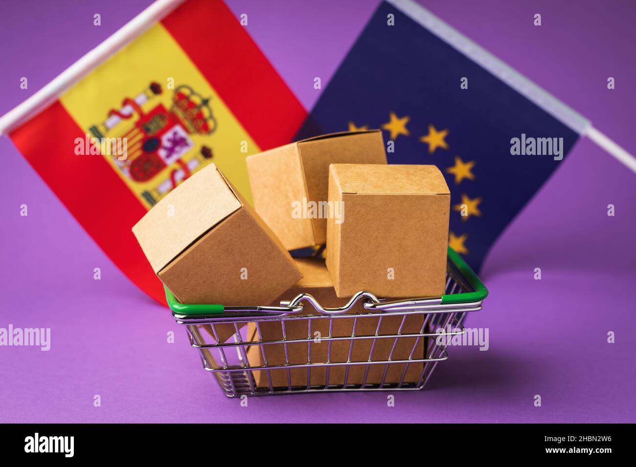 Scatole di cartone in un cestino alimentare e bandiere su sfondo colorato, concetto sulla distribuzione di merci spagnole nell'Unione europea Foto Stock