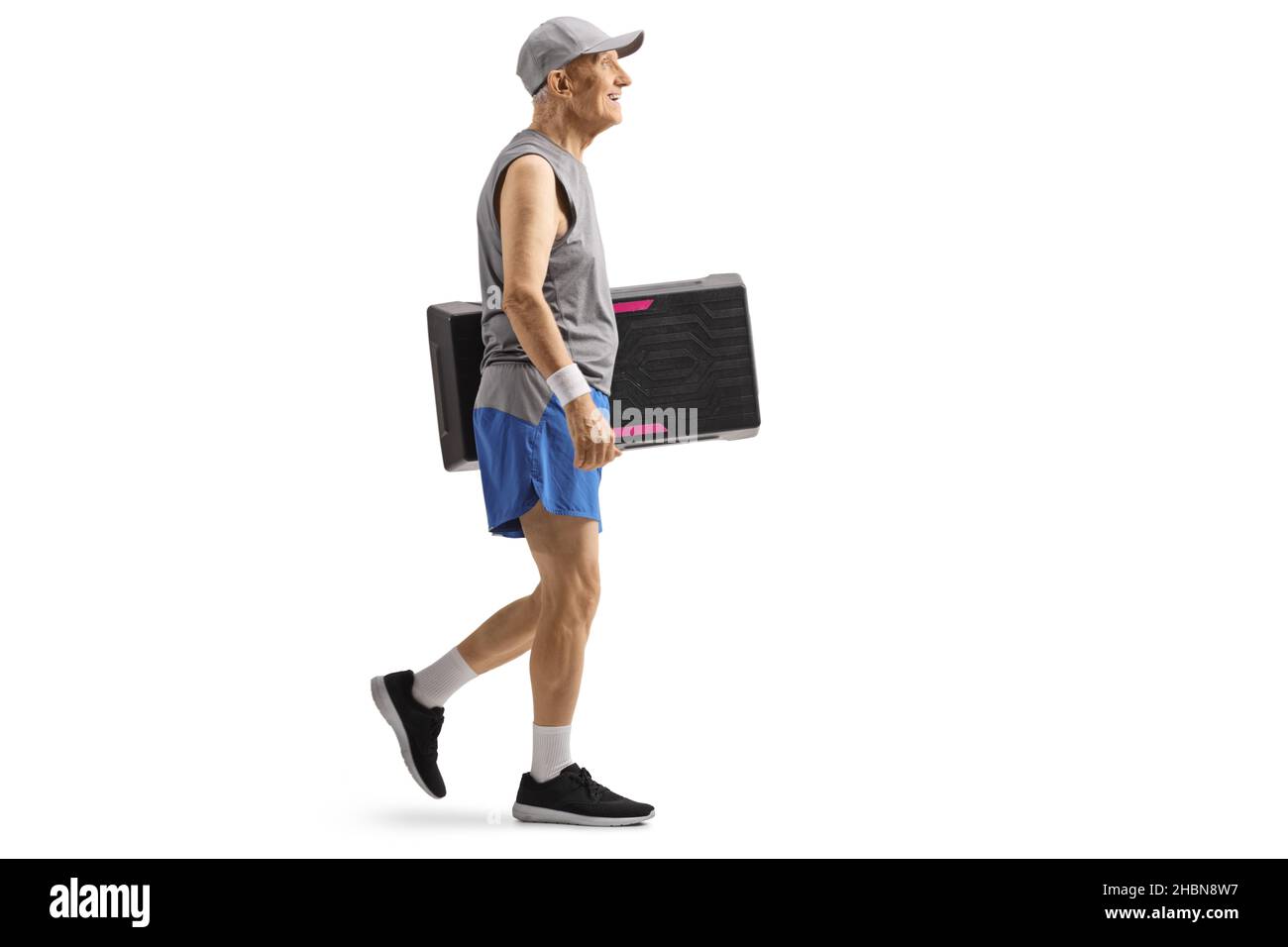 Scatto a profilo completo di un uomo anziano in abbigliamento sportivo che cammina e trasporta una piattaforma aerobica step isolata su sfondo bianco Foto Stock