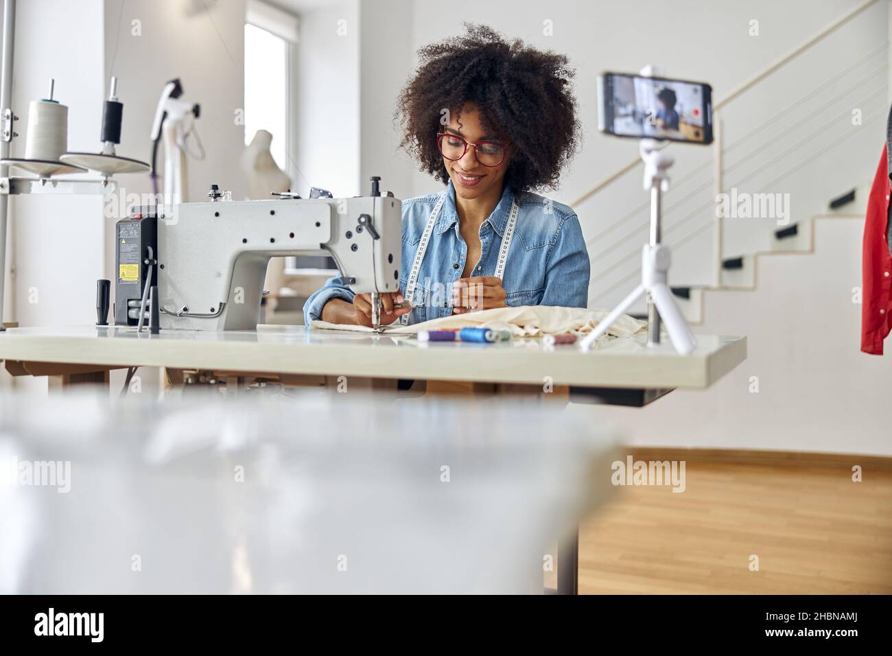 Positivo Afro-American dressmaker tira tutorial sul posto di lavoro con la macchina da cucire in studio Foto Stock