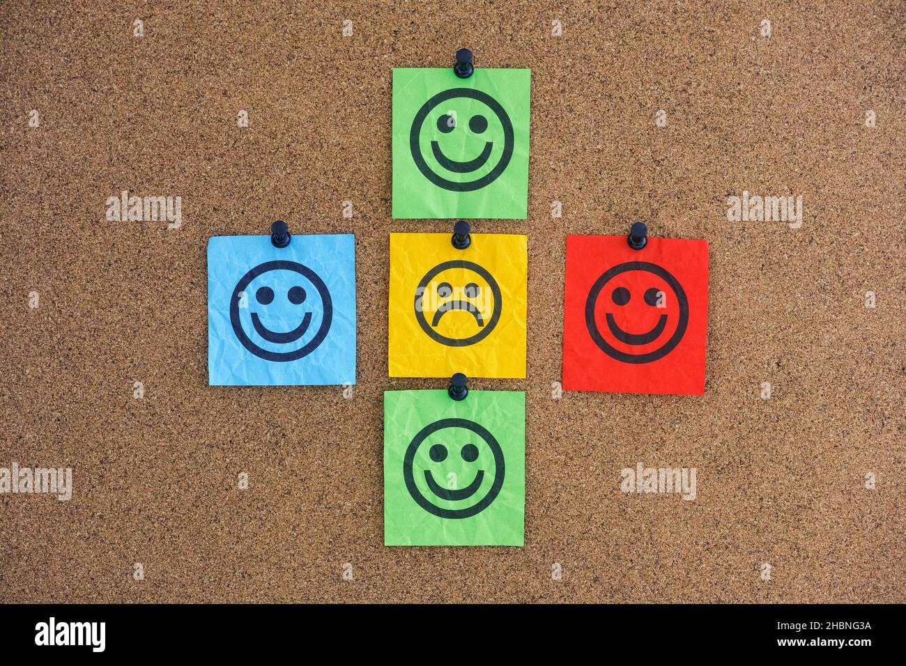 Una nota di carta con un volto triste con volti felici intorno ad esso su un bordo del corkboard. Concetto di rifiuto sociale. Primo piano. Foto Stock