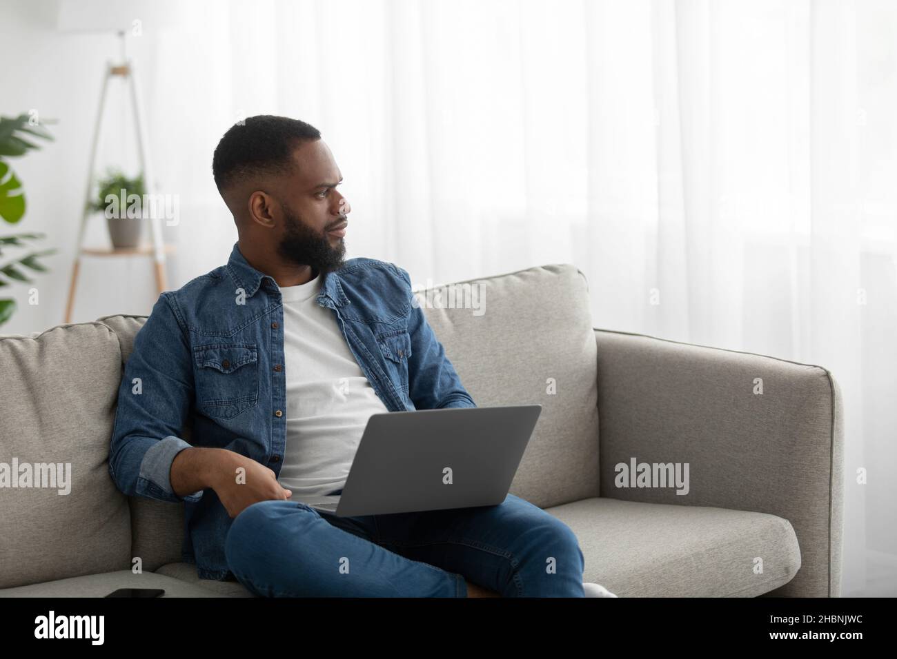 Serio concentrato giovane nero barbuto lavoro su computer portatile sul divano interno della stanza, guardare la finestra Foto Stock
