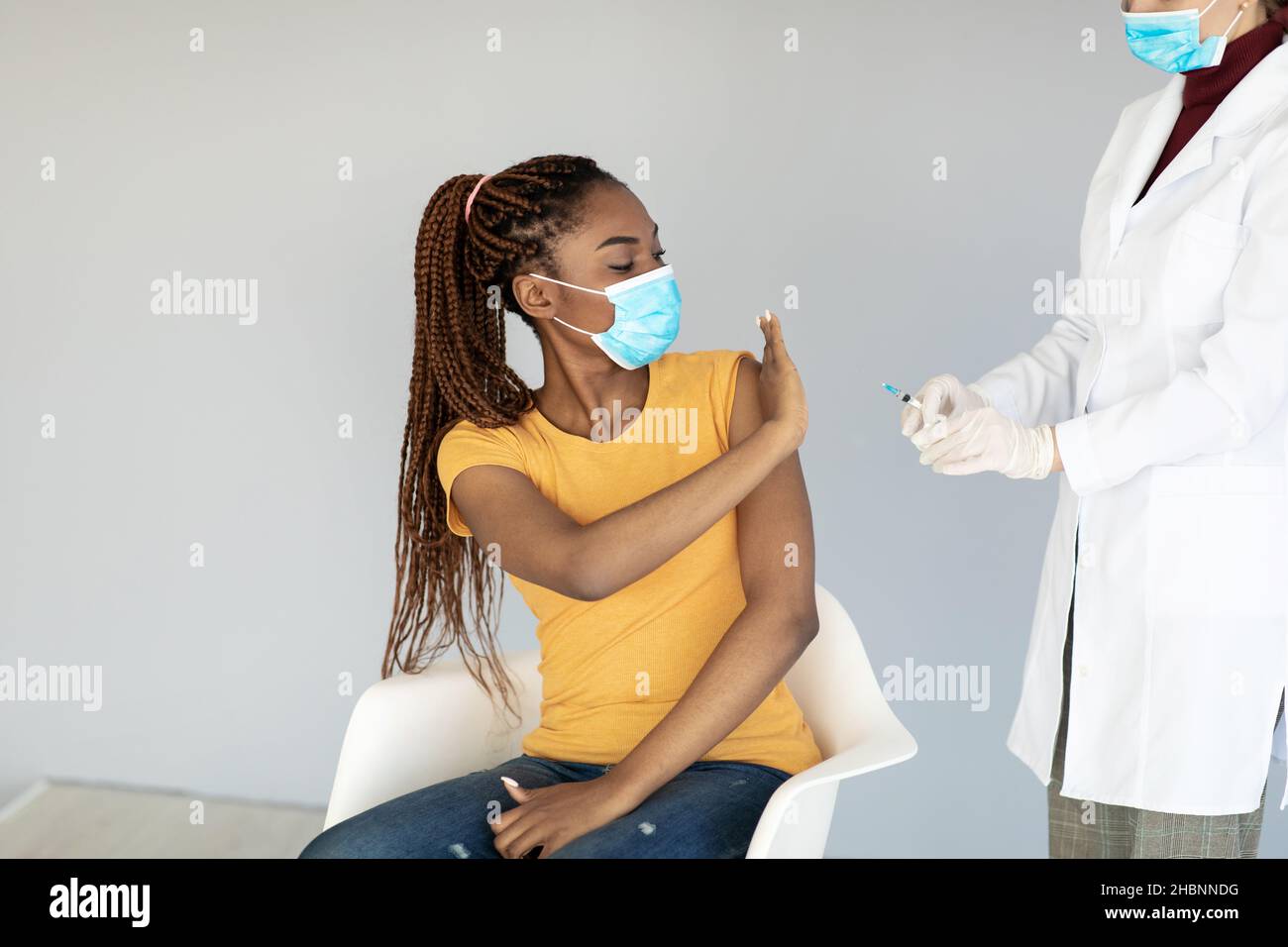 Giovane donna nera in maschera facciale usa e getta che impedisce al medico di fare l'iniezione di vaccino del coronavirus su sfondo grigio Foto Stock