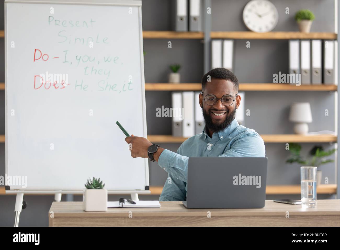 Allegro giovane insegnante di tipo nero con pc mostra su lavagna con regole di lingua inglese Foto Stock