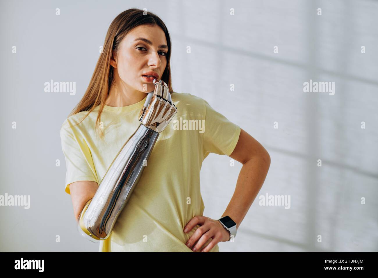 Donna piuttosto premurosa con braccio protesico bionico in metallo lucido in posa t-shirt per macchina fotografica in piedi a parete in primo piano stanza Foto Stock