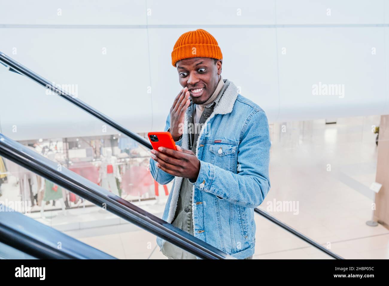 Emozionale felice africano-americano ragazzo in elegante vestito guarda il telefono cellulare che va su scala mobile nel centro commerciale vicino vista Foto Stock