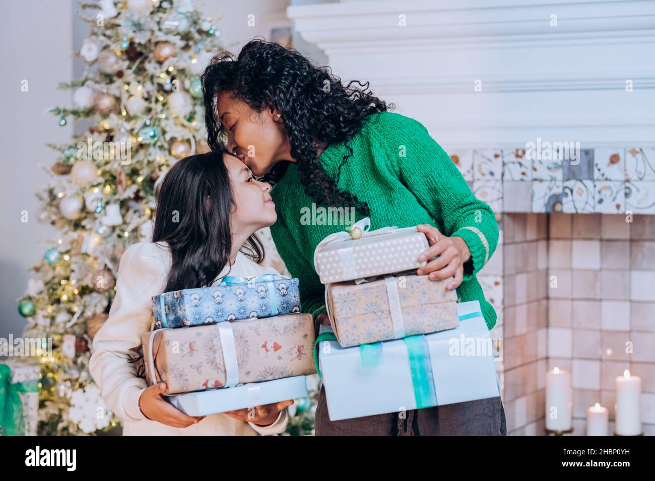 Mamma africana americana in maglione verde e figlia con capelli lunghi scuro stand con regali di natale vicino all'albero di natale, madre bacia figlia sulla fronte Foto Stock