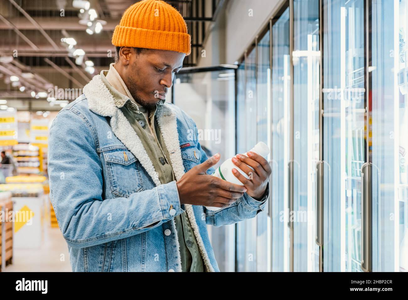 Il ragazzo afro-americano positivo in giacca di denim e cappello arancione prende la bottiglia di latte dalla scatola del frigorifero in vista laterale del moderno supermercato Foto Stock