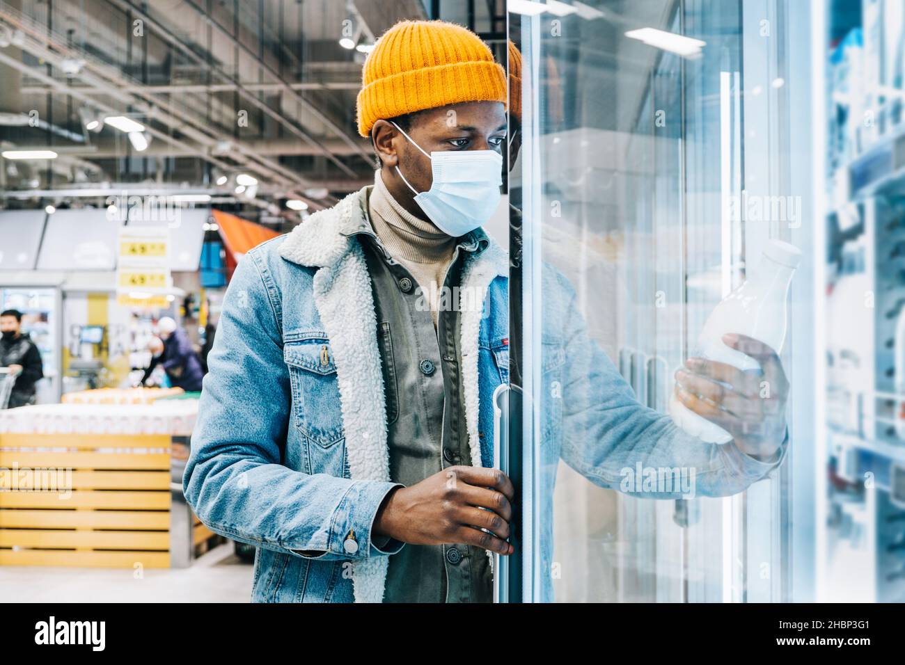Il giovane uomo afro-americano in giacca denim con maschera usa e getta prende la bottiglia di latte dalla scatola del frigorifero nella moderna sala del supermercato Foto Stock
