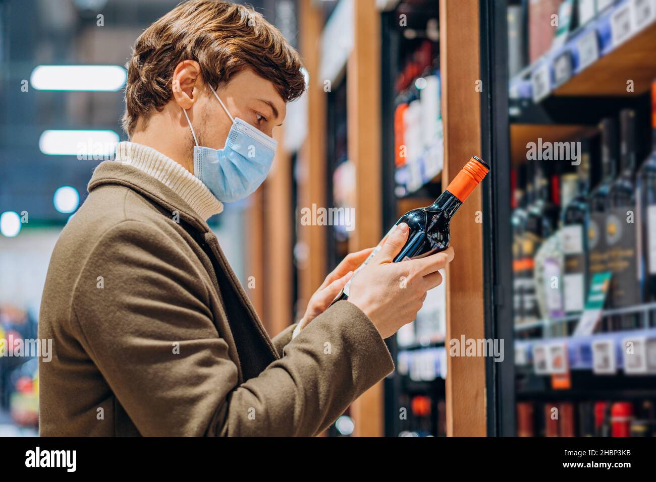 l'uomo di successo in un cappotto con maschera legge l'etichetta su una bottiglia di vino in un moderno supermercato, sceglie una bevanda alcolica, studia la composizione Foto Stock