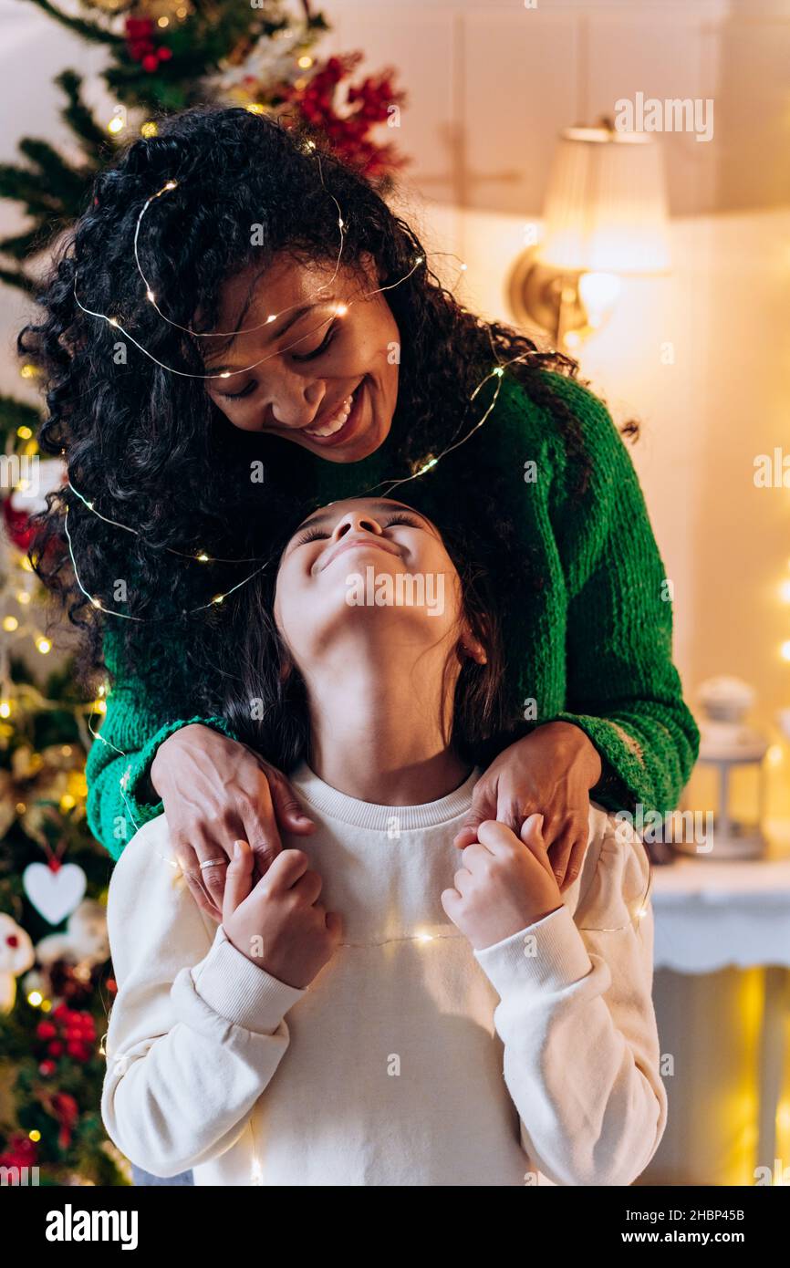 La ragazza della bruna e la madre afroamericana lunga ricci si posa con la ghirlanda illuminante sulla testa e sorrida felicemente contro l'albero di Natale Foto Stock