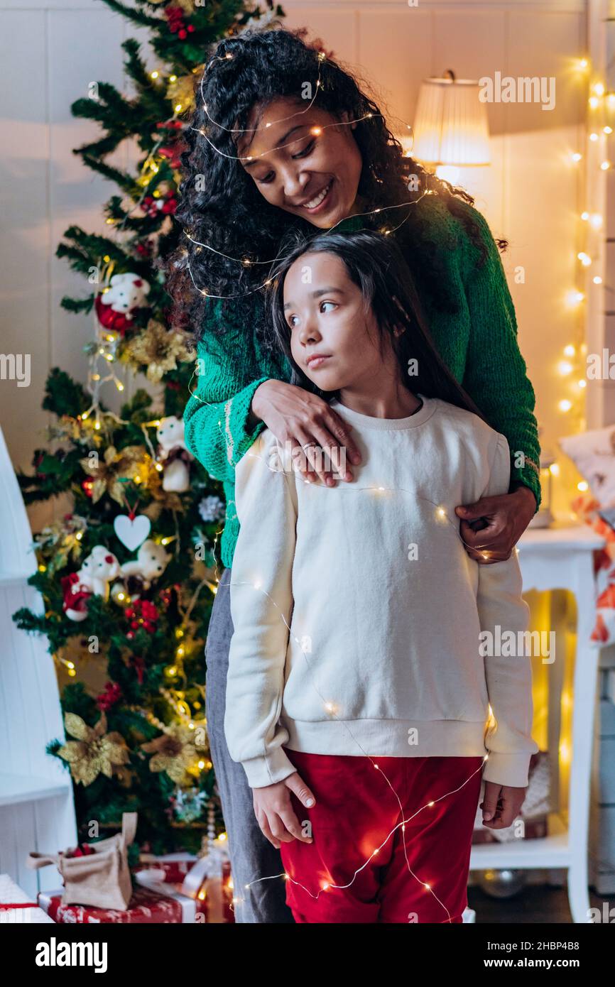 La ragazza della bruna e la madre afroamericana lunga ricci si posa con la ghirlanda illuminante sulla testa e sorrida felicemente contro l'albero di Natale Foto Stock