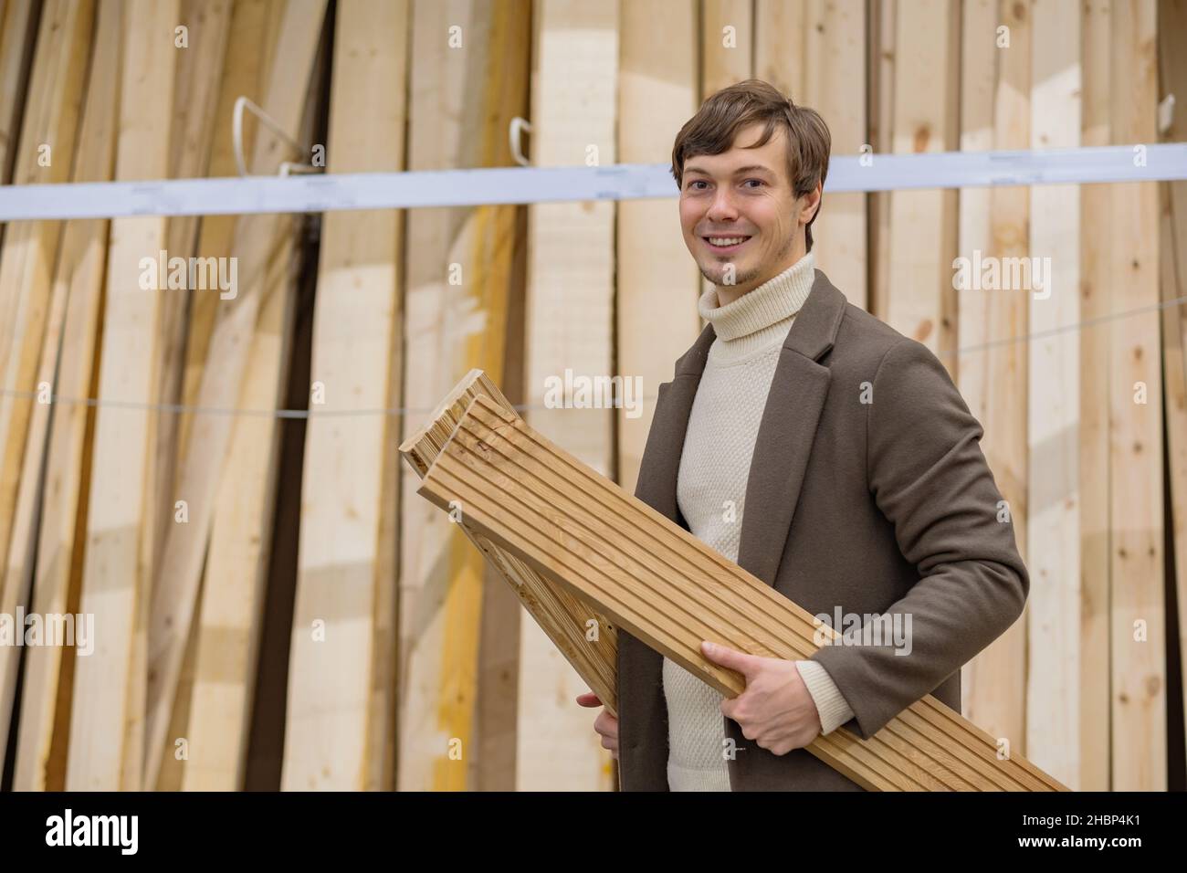 ritratto di un uomo di successo in un cappotto marrone in un negozio di ferramenta guardando la macchina fotografica con assi di legno nelle sue mani Foto Stock