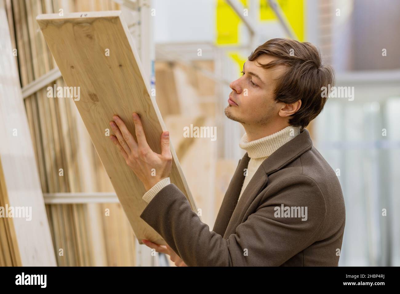 l'uomo riuscito in un cappotto marrone seleziona con cura le tavole di legno per la costruzione in un negozio di riparazione Foto Stock