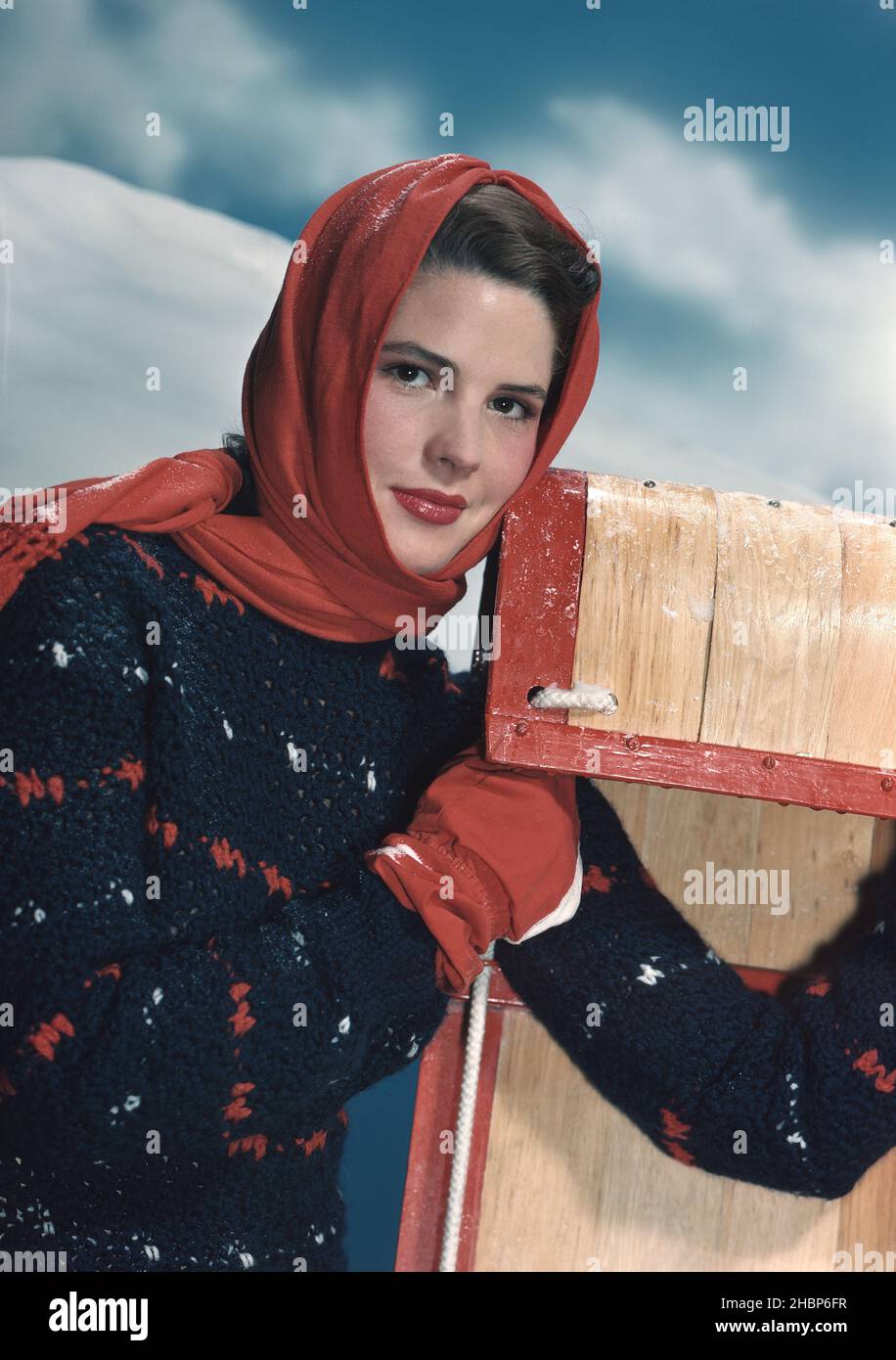 Teen girl in blue print sweater and red hood standing in snow holding a toboggan Foto Stock