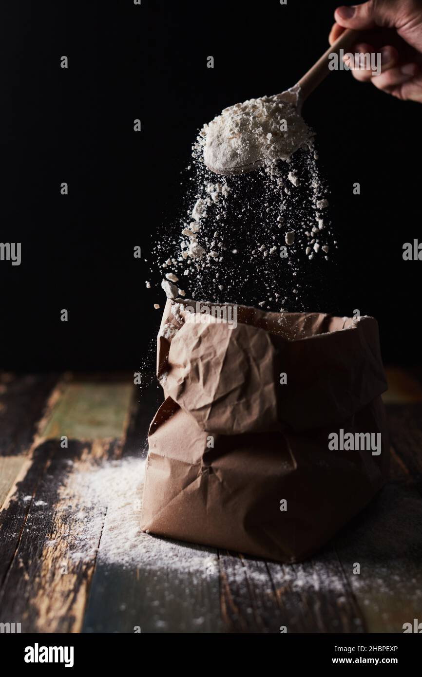 Farina di grano in un pacchetto di mestiere. Movimento di congelamento, cucchiaio sfondo scuro, spolverare la farina. Vista frontale. Foto Stock