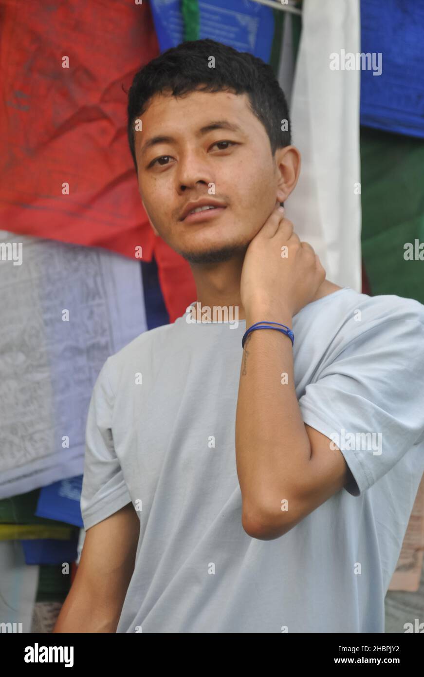 Foto di un ragazzo asiatico in posa con mano dietro il collo guardando la macchina fotografica, in piedi contro le bandiere buddiste di preghiera Foto Stock