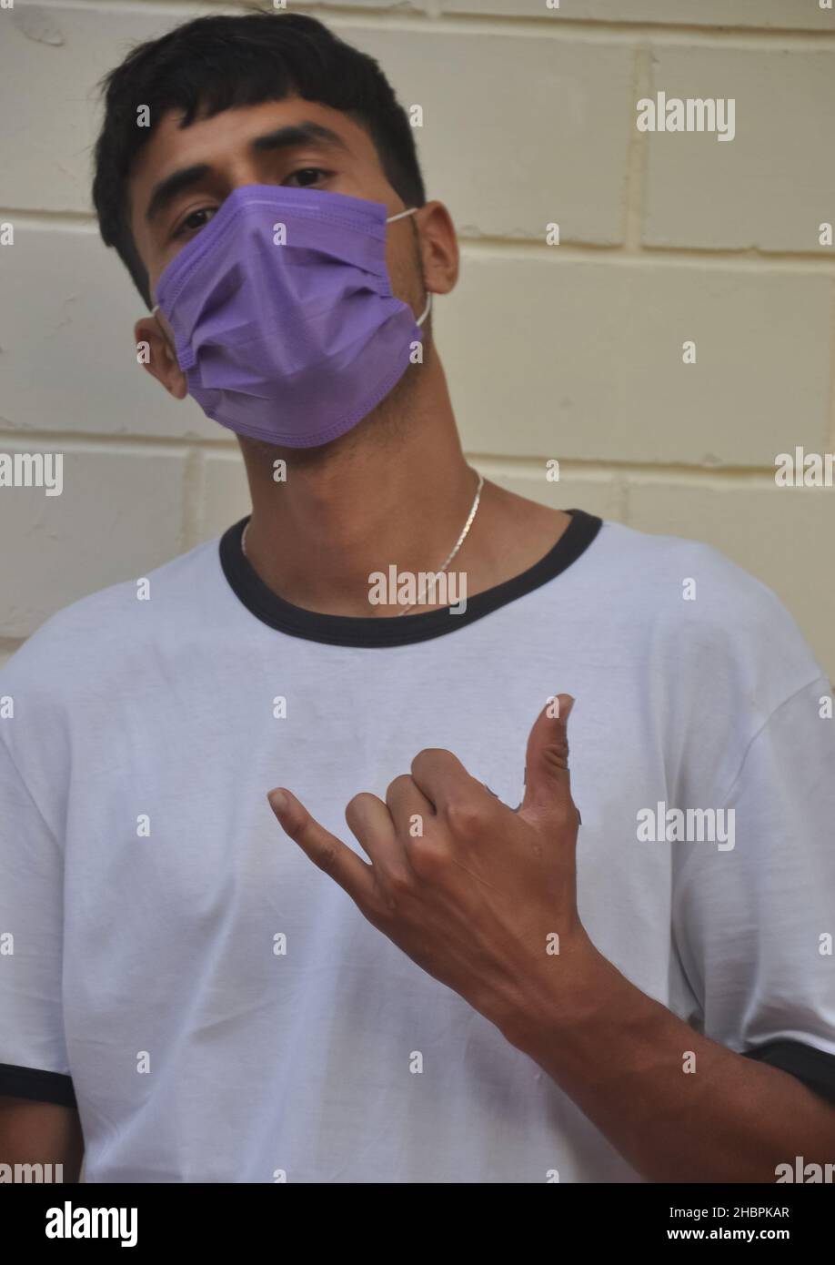 Un ragazzo giovane che fa segno shaka con la sua mano, indossare maschera facciale con guardare la macchina fotografica mentre contro la parete bianca Foto Stock