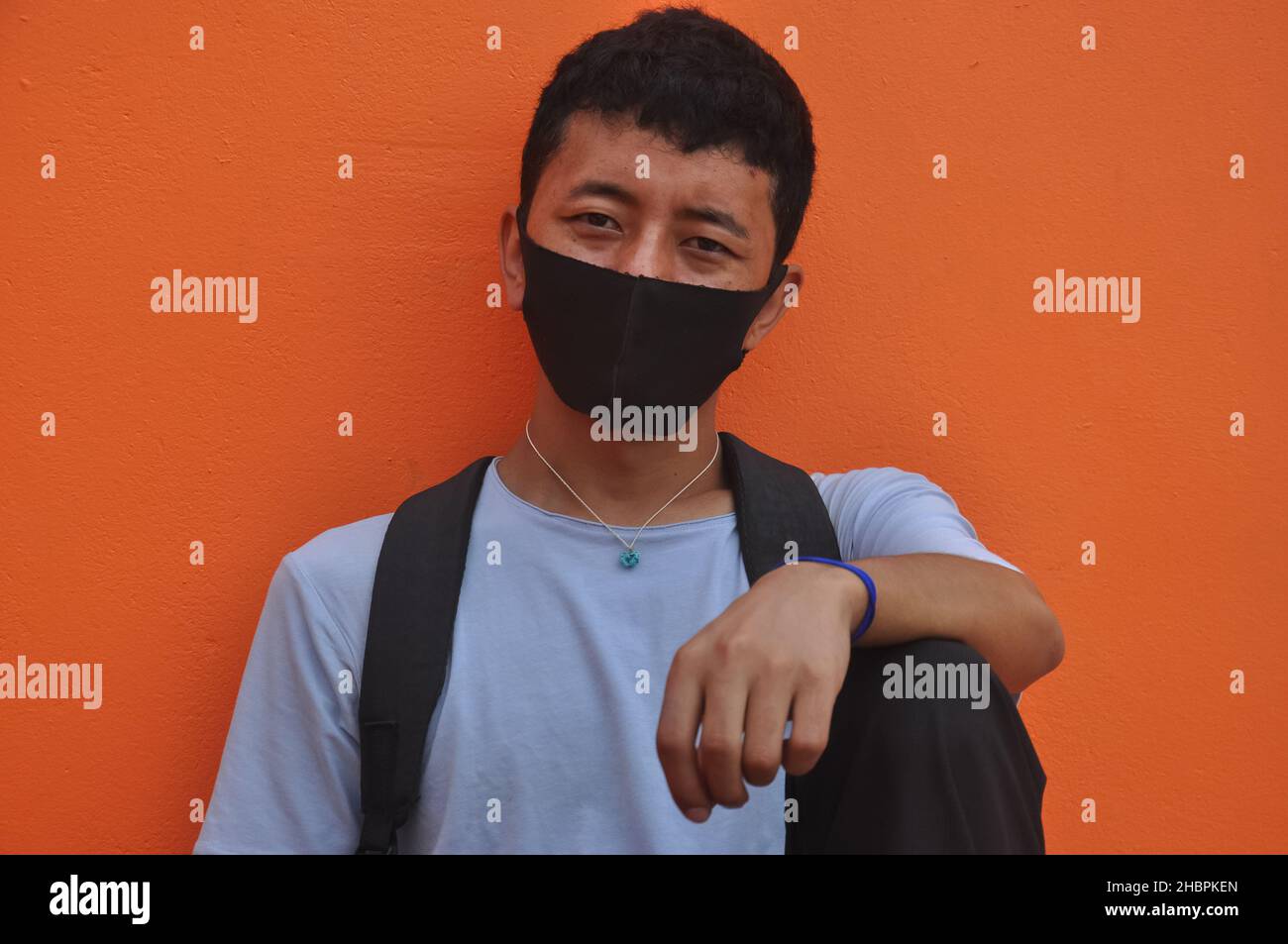 Vista frontale di un giovane asiatico che indossa maschera facciale con guardare la macchina fotografica seduta su sfondo arancione parete con la mano sul ginocchio Foto Stock
