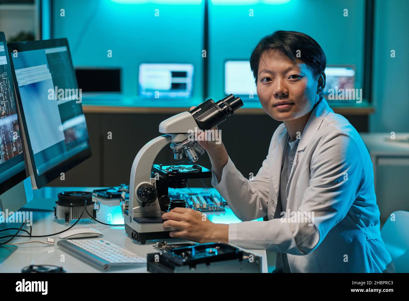 Giovane ricercatore asiatico in whitecoat lavorando con il microscopio e guardando voi mentre seduti sul posto di lavoro in laboratorio Foto Stock