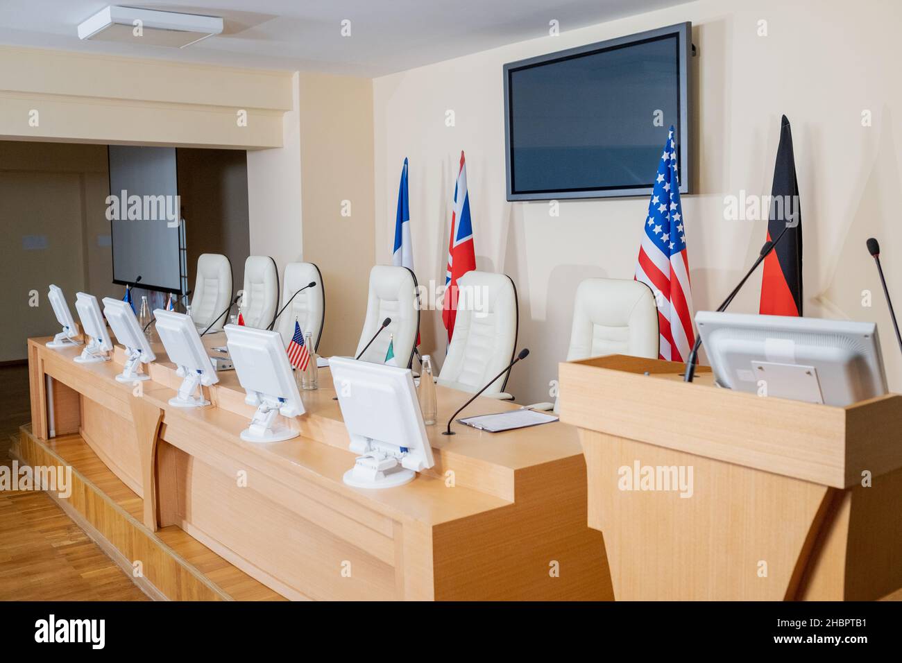 Sala conferenze vuota con fila di microfoni, monitor per computer e bandiere su tavolo lungo, tribuna e poltrone Foto Stock