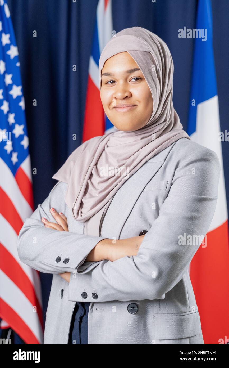 Giovane uomo politico musulmano ben vestito in hijab ed elegante vestito in piedi davanti alla macchina fotografica sullo sfondo di diverse bandiere Foto Stock