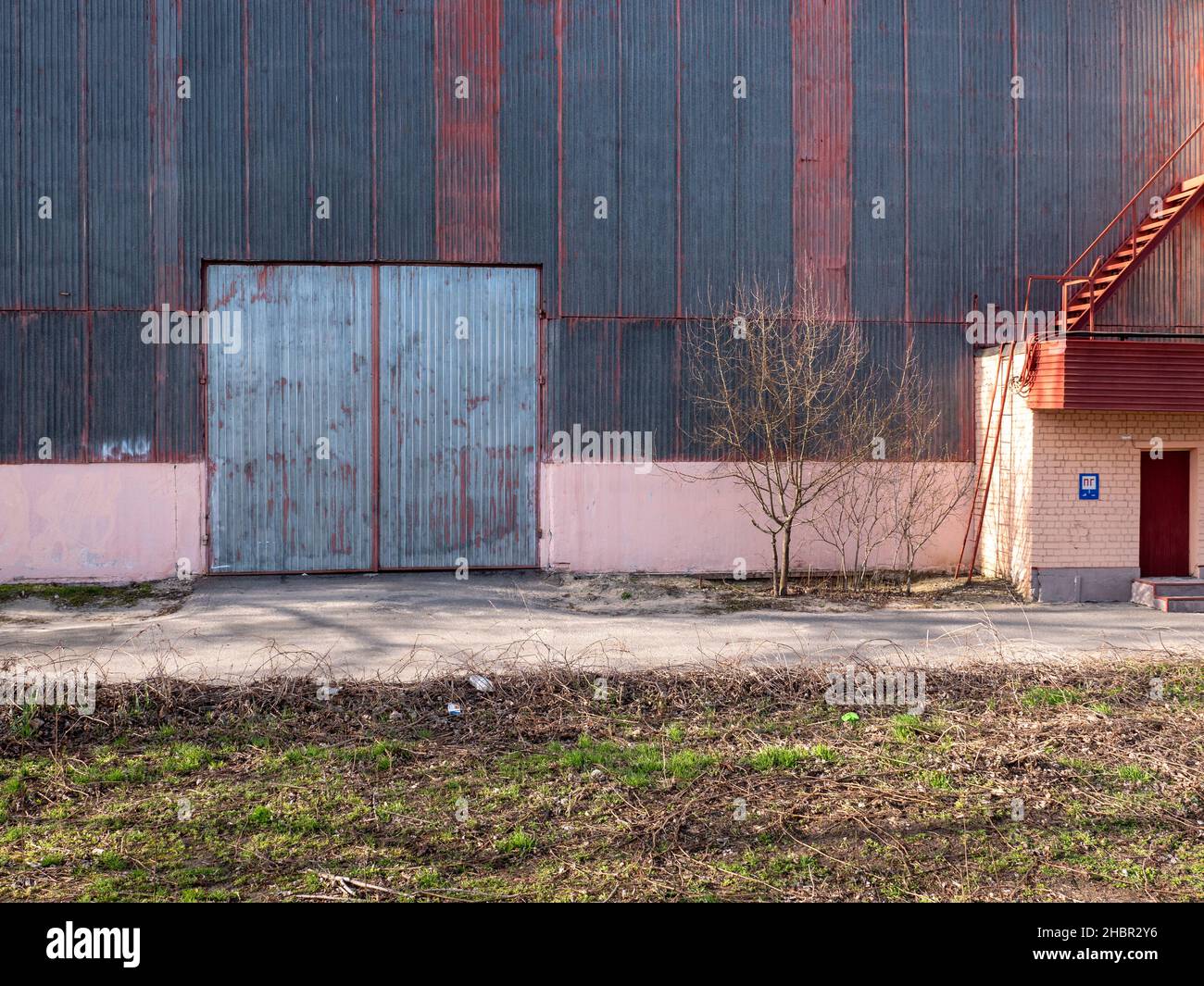Vecchio cancello di metallo alto scanalatura con vernice grigia peeling del vecchio magazzino con vecchia facciata rivestita di metallo in una fredda giornata di sole. Porta e scala sul tetto. Foto Stock