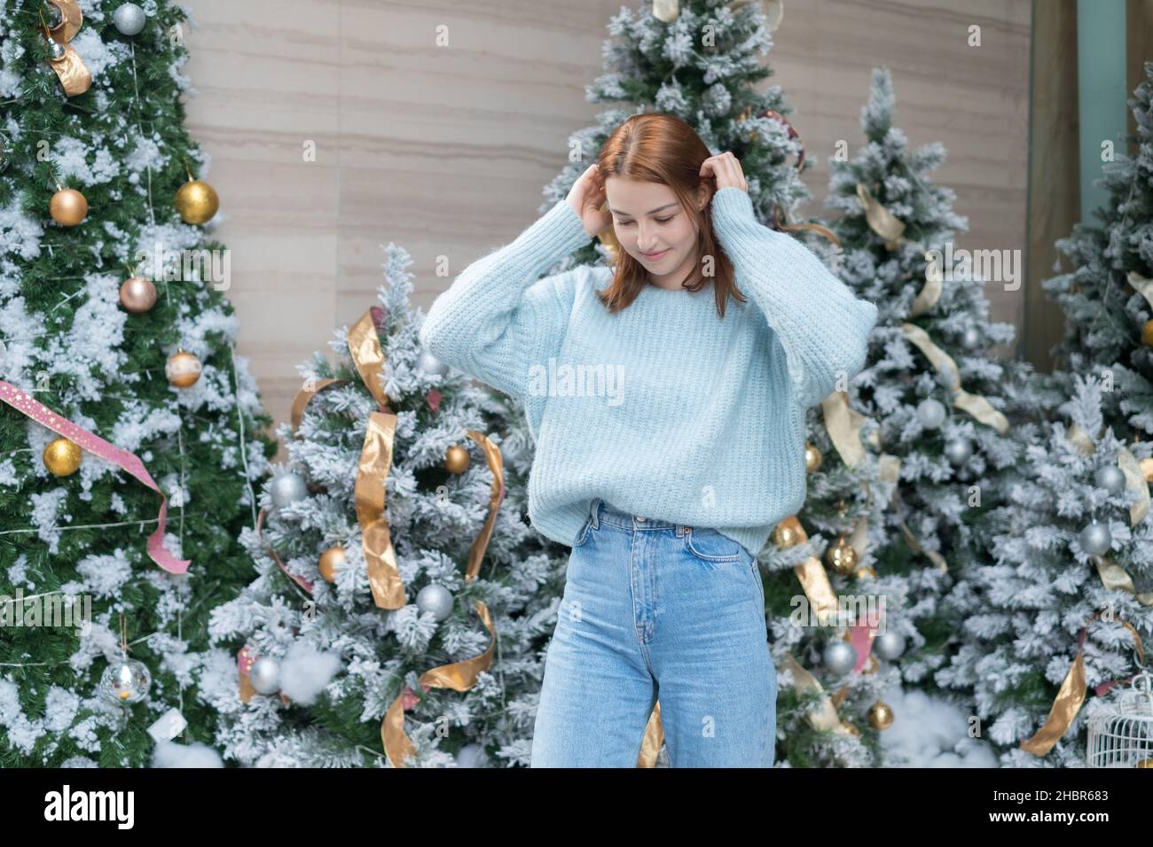 Ritratto di Capodanno di bella ragazza caucasica sorridente in lana accogliente caldo azzurro maglione in piedi vicino decorato albero di Natale. Persona felice Foto Stock