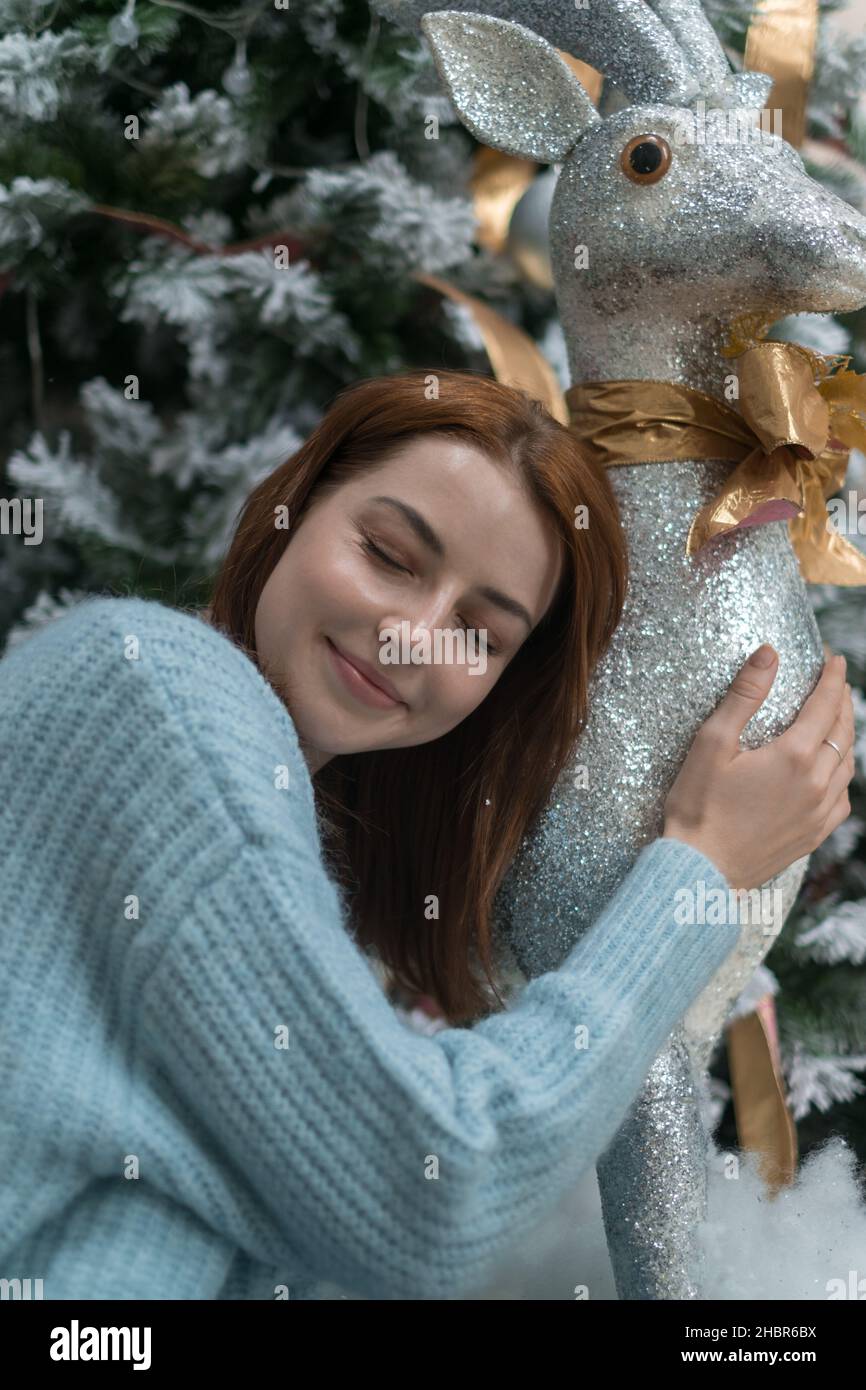 Ritratto di Capodanno di adorabile ragazza caucasica sorridente in lana caldo blu maglione. Ragazza si siede vicino l'albero di Natale e coccole giocattolo renna Foto Stock