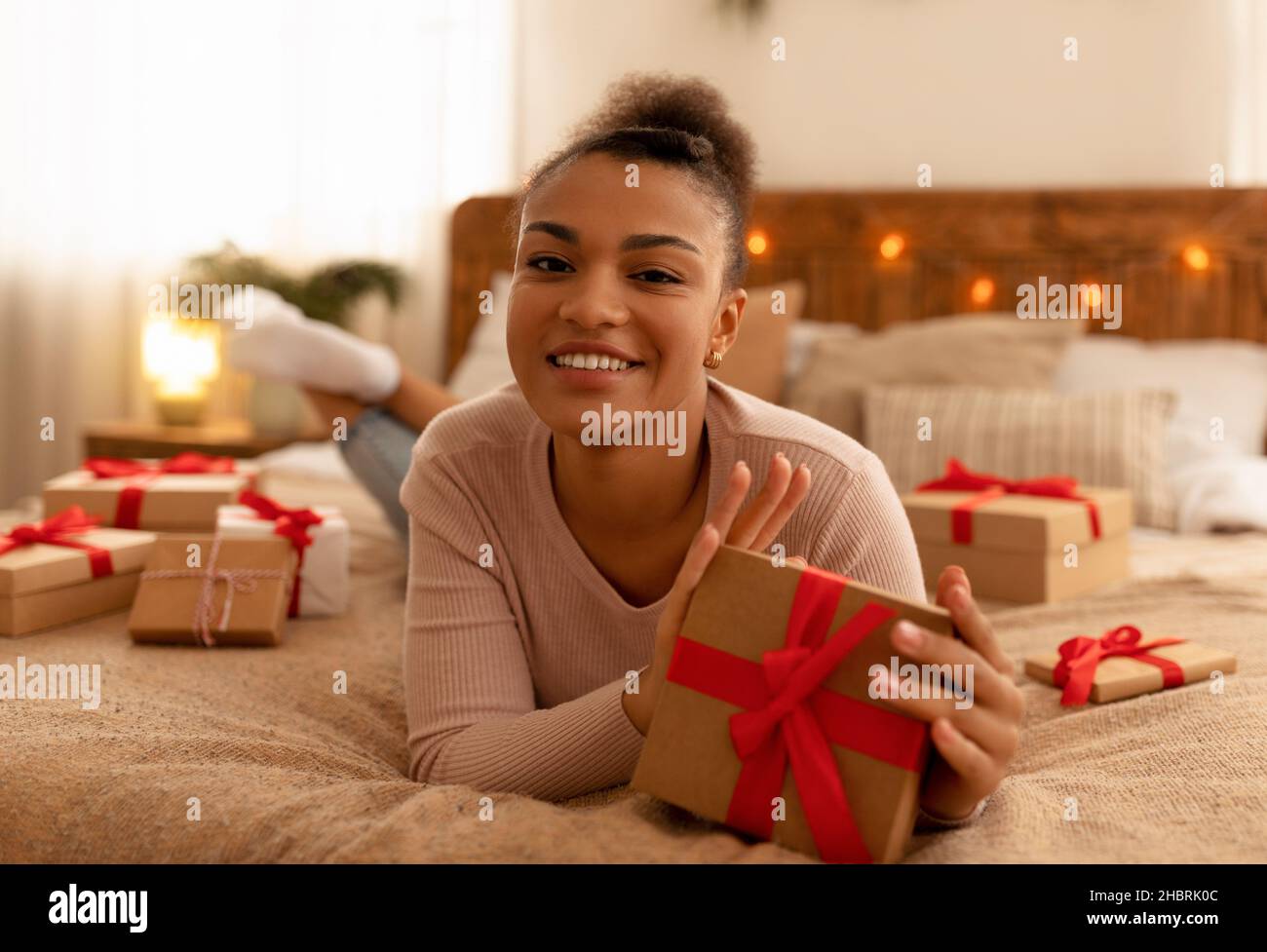 Felice giovane afroamericana signora sdraiata a letto e tenendo in mano la scatola regalo di Natale, sorridendo alla macchina fotografica Foto Stock