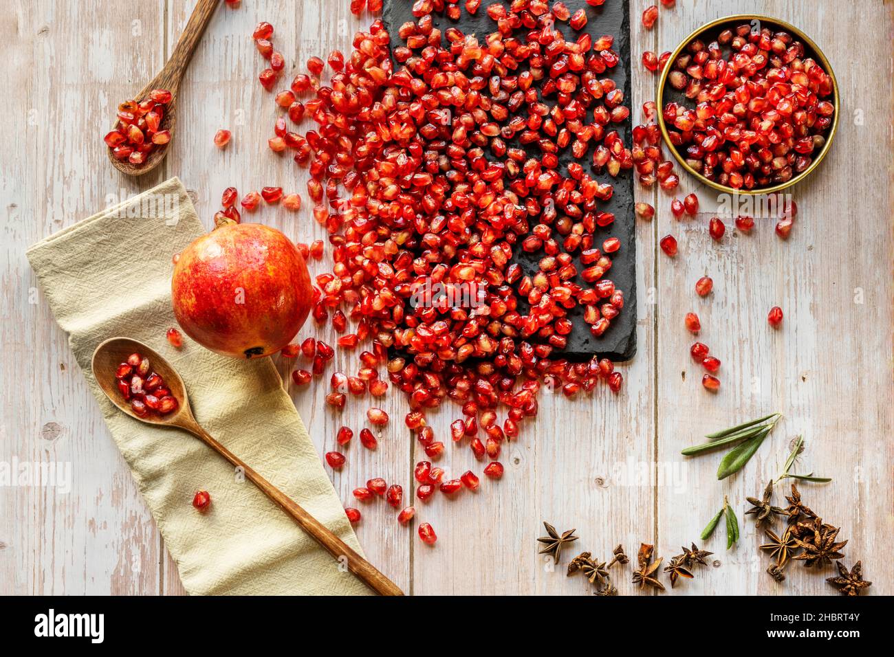 Immagine dall'alto di un gran numero di grani di melograno rosso con cucchiai di legno, anice, piatti d'annata e tessuto giallo Foto Stock