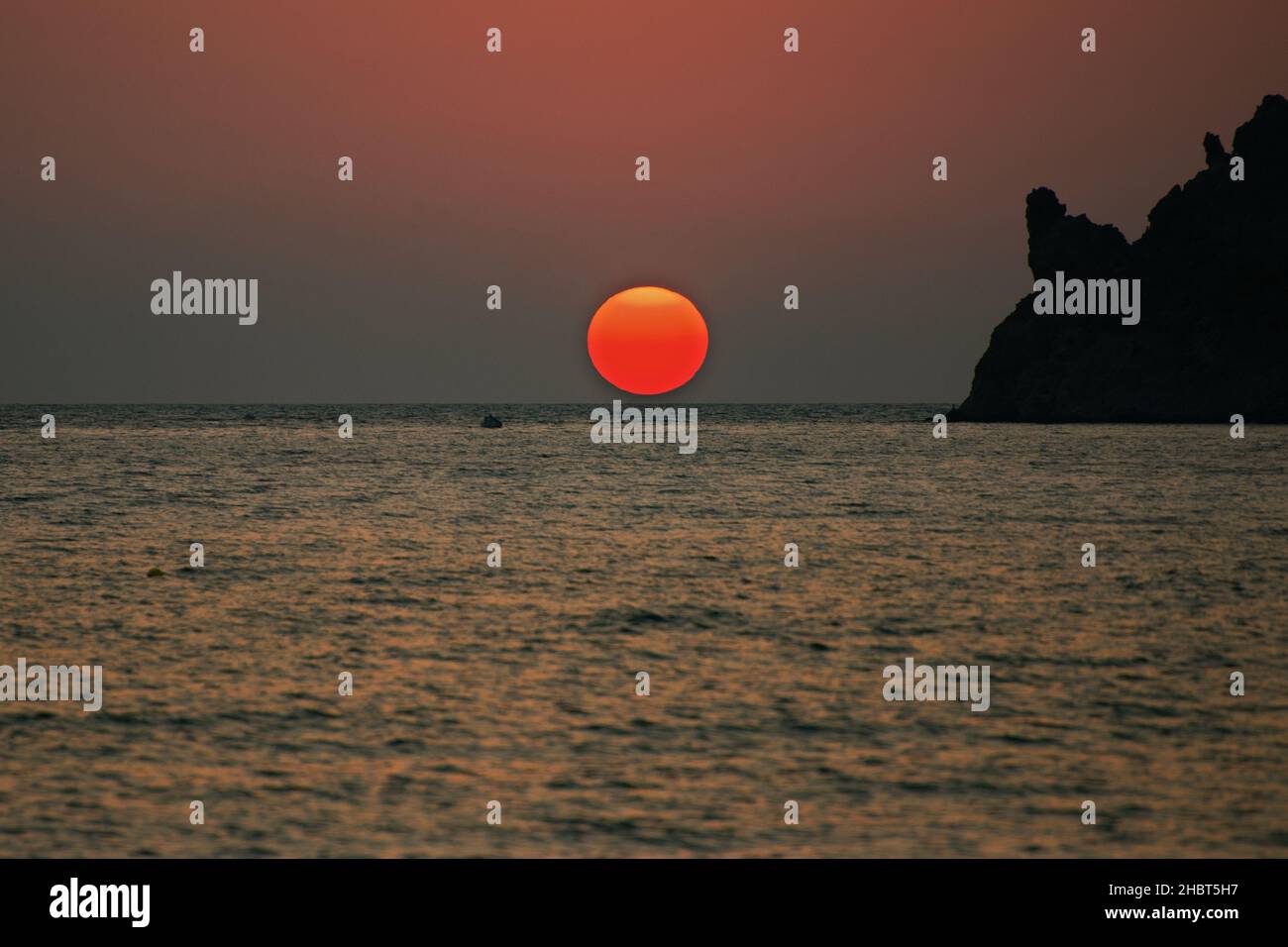 Grande disco solare rosso, superficie ondulata del mare, piccola barca all'orizzonte e parte della silhouette rocciosa al tramonto. LIMNOS, Grecia. Foto Stock