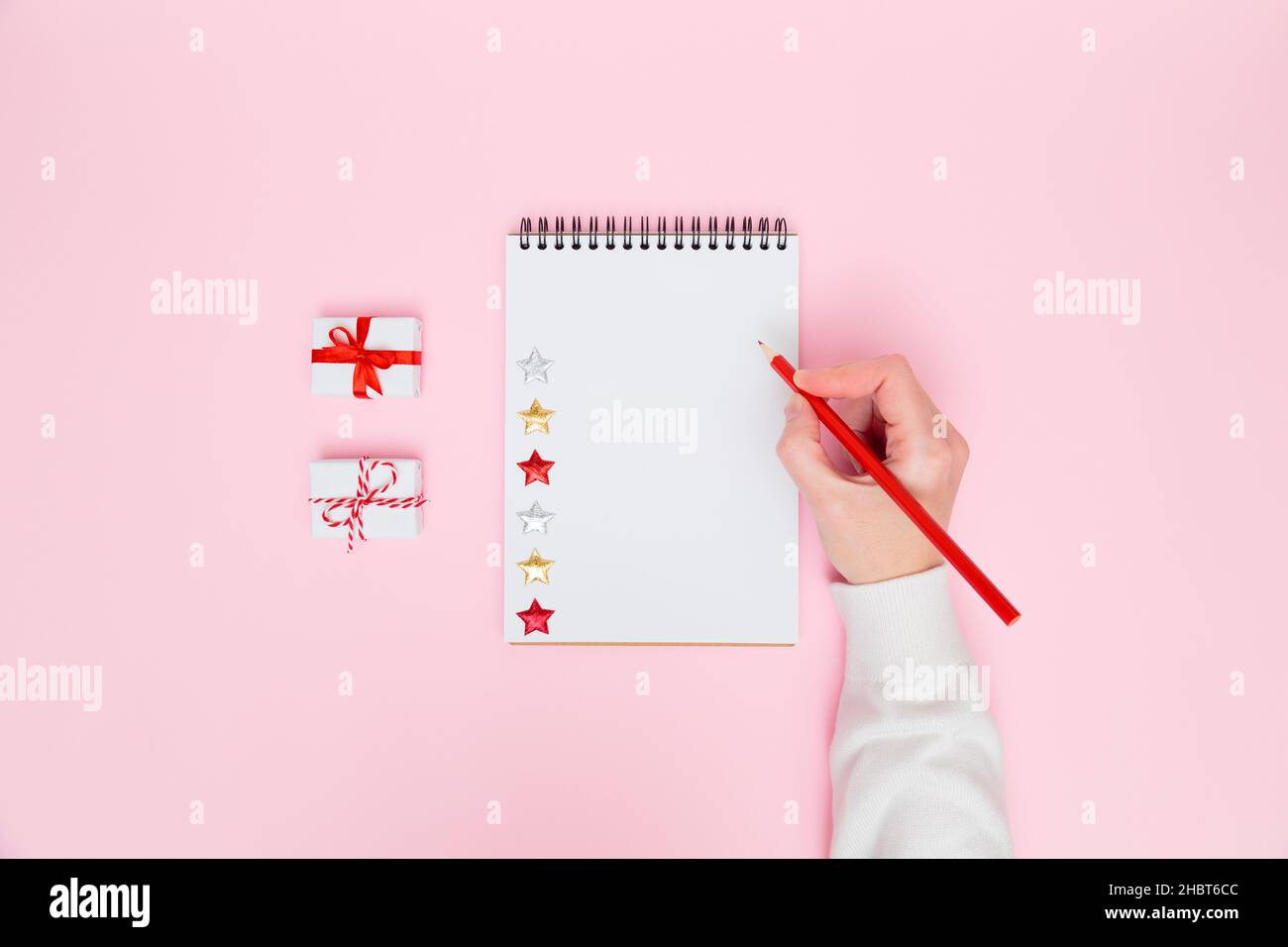 Sfondo accogliente Natale. Scrittura femminile a mano in taccuino bianco con scatole regalo bianche con archi rossi su rosa pastello. Pianificazione del nuovo anno, obiettivi, da fare l Foto Stock