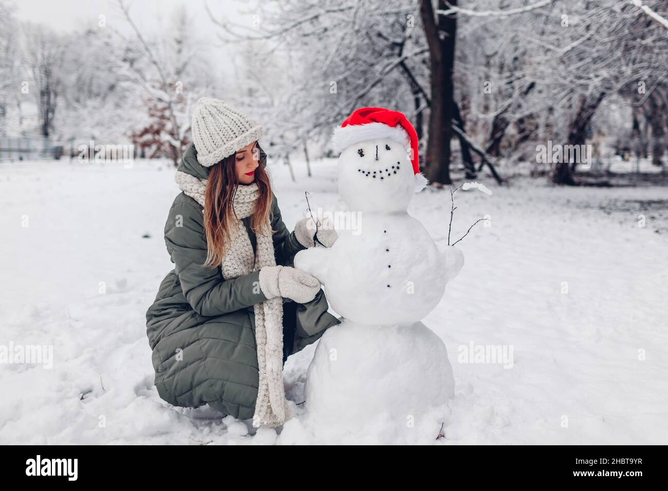 La ragazza fa il pupazzo di neve all'aperto nel parco invernale nevoso vestendolo in Santa Hat, sciarpa ed attaccando i rami nelle mani. Attività divertenti di Natale Foto Stock
