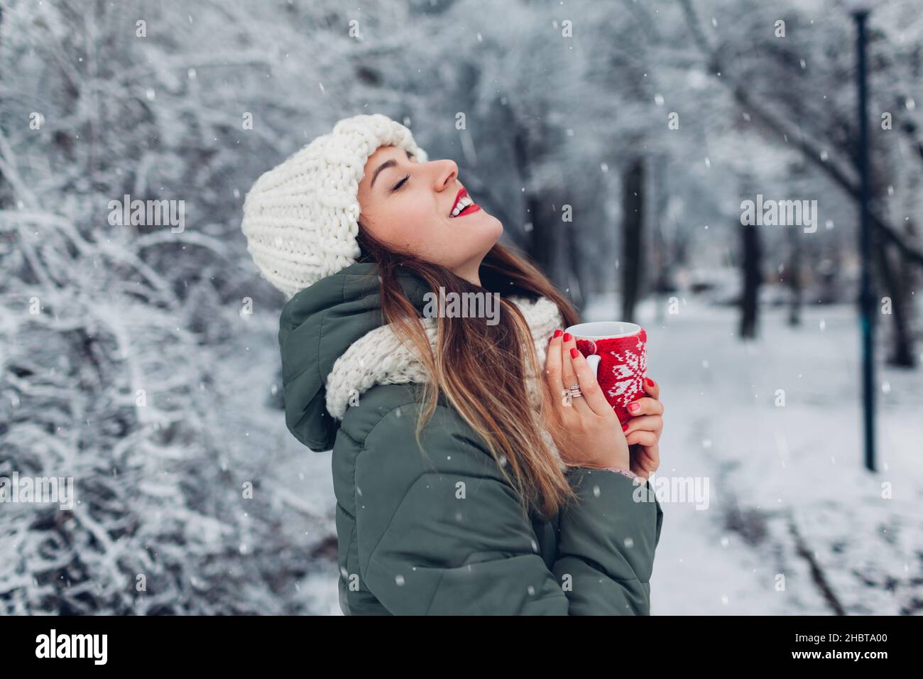 Giovane donna che beve tè caldo o caffè che tiene il pallone da vuoto in nevoso parco invernale che respira liberamente. Tazza vestita in una custodia di Natale in maglia rossa Foto Stock
