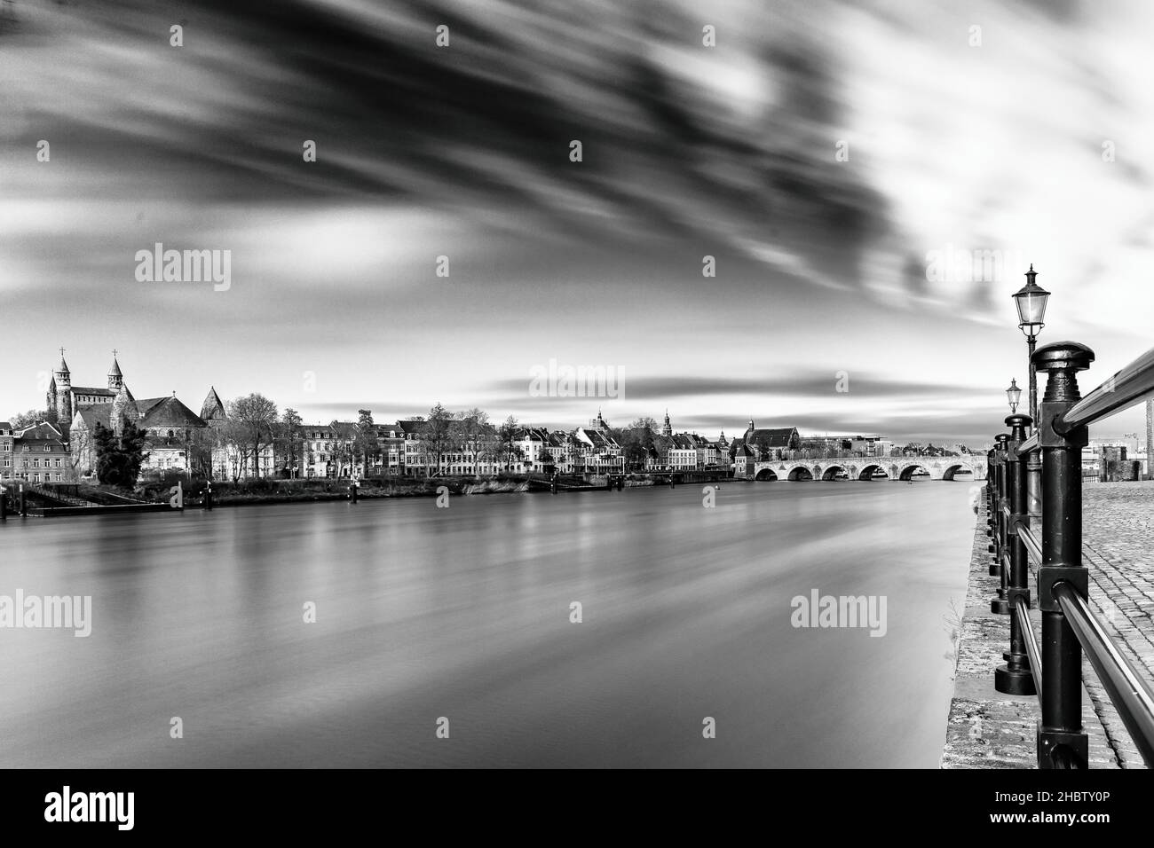 Lunga esposizione in bianco e nero del centro di Maastricht con una vista sulle acque setose del fiume Meuse e un cielo drammatico pieno di nuvole in movimento. Foto Stock
