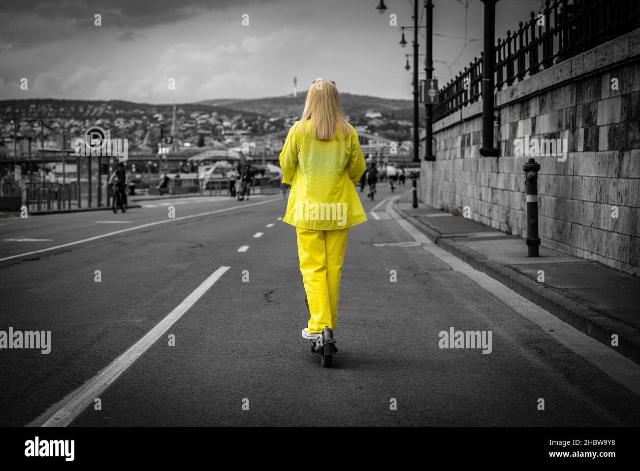 Donna bionda in abiti gialli utilizzando uno scooter elettrico sul molo del Danubio a Budapest, girato da dietro, giallo su sfondo bianco e nero Foto Stock