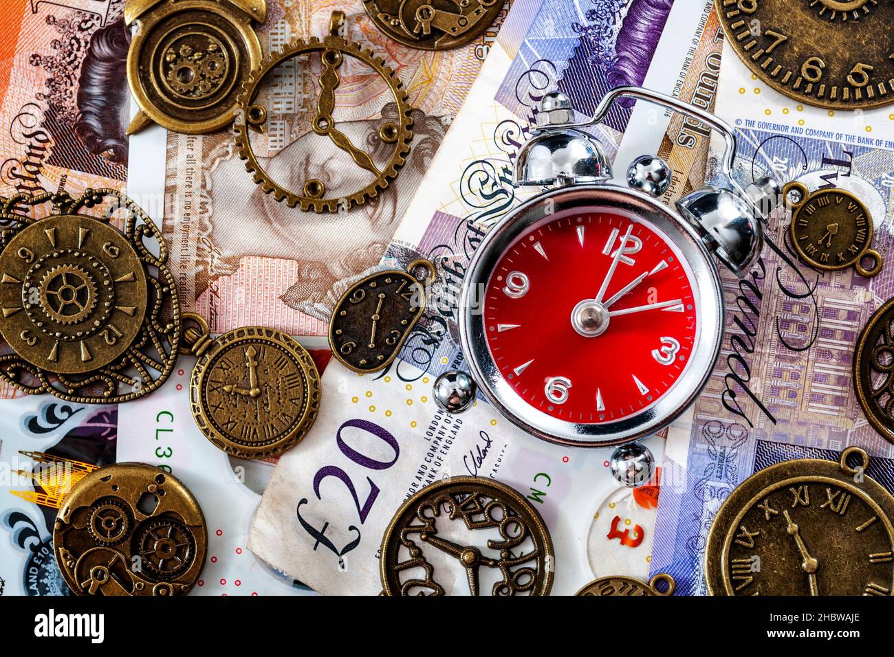 Il tempo è concetto di denaro con piatto di sveglia su diverse banconote o banconote inglesi di denominazione e circondato da orologi in metallo Foto Stock