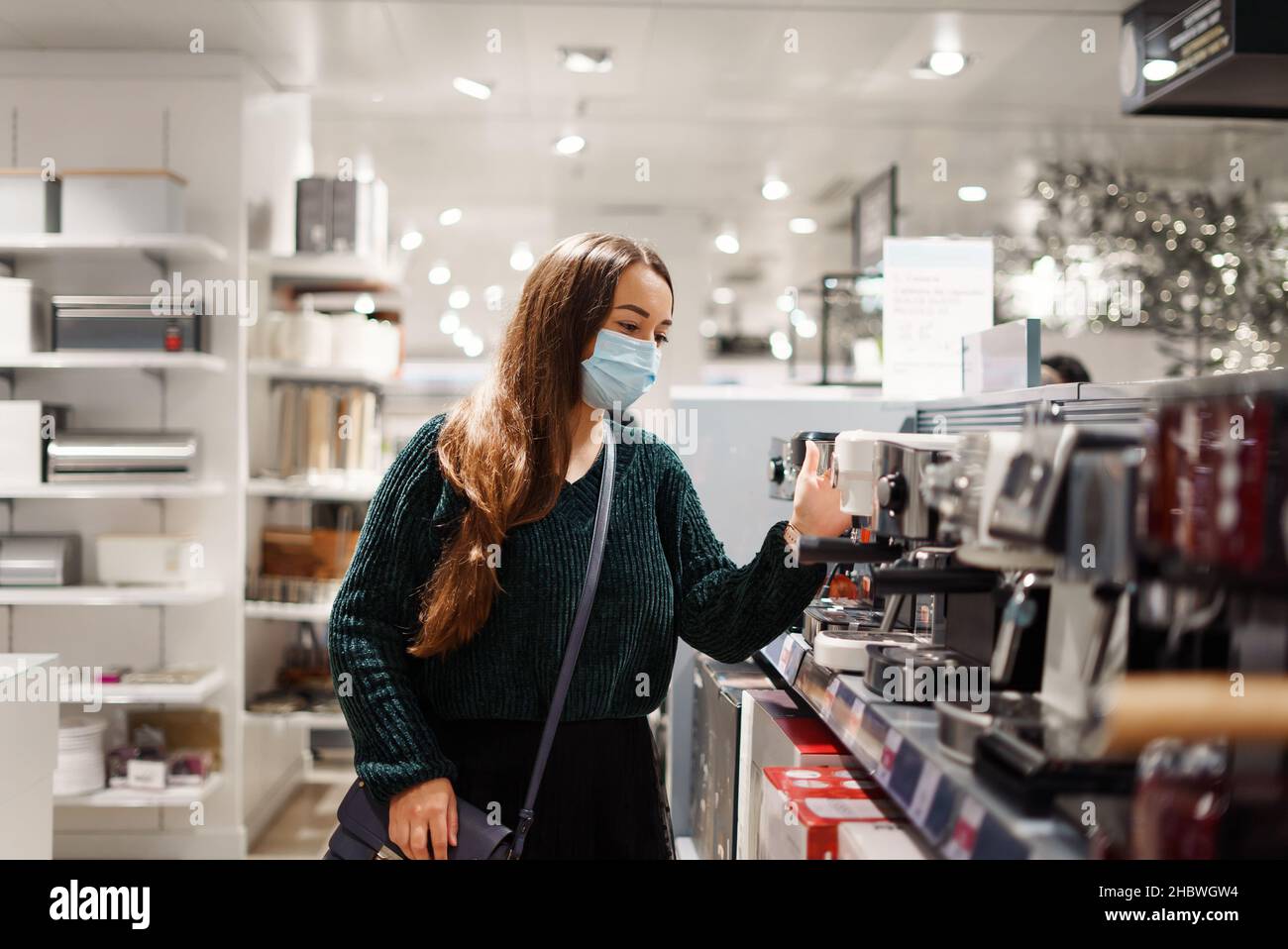 La giovane donna in maschera facciale sceglie la macchina da caffè in negozio, considerando i prezzi, confrontando le specifiche tecniche e i marchi di fabbrica per gustare fragrante tazza di espresso al giorno. Shopping durante covid19 pandemic Foto Stock