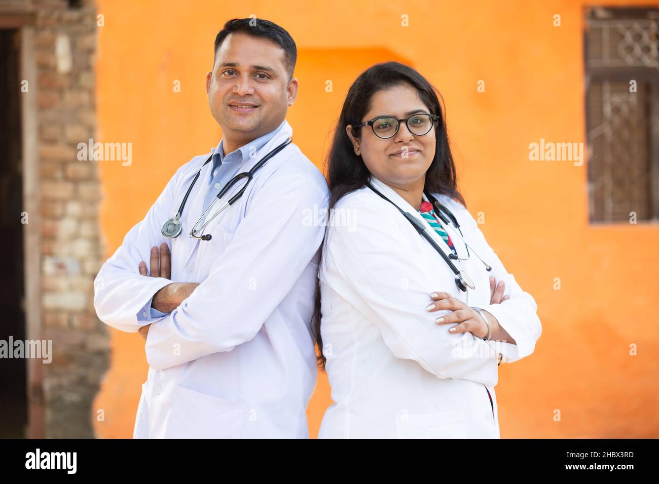Ritratto di giovane uomo indiano sorridente e femmina medico indossando stetoscopio Croce Arms in piedi guardando fotocamera con emozioni positive contro orang Foto Stock
