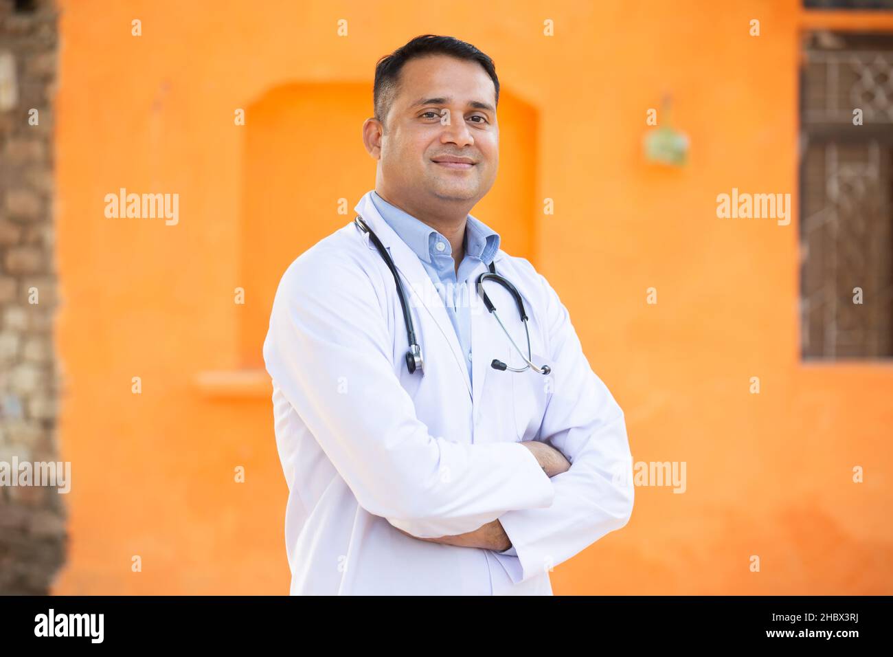 Ritratto di giovane uomo indiano sorridente medico indossando stetoscopio Standing Cross Arms, sta guardando la macchina fotografica con emozioni positive contro Orange ba Foto Stock