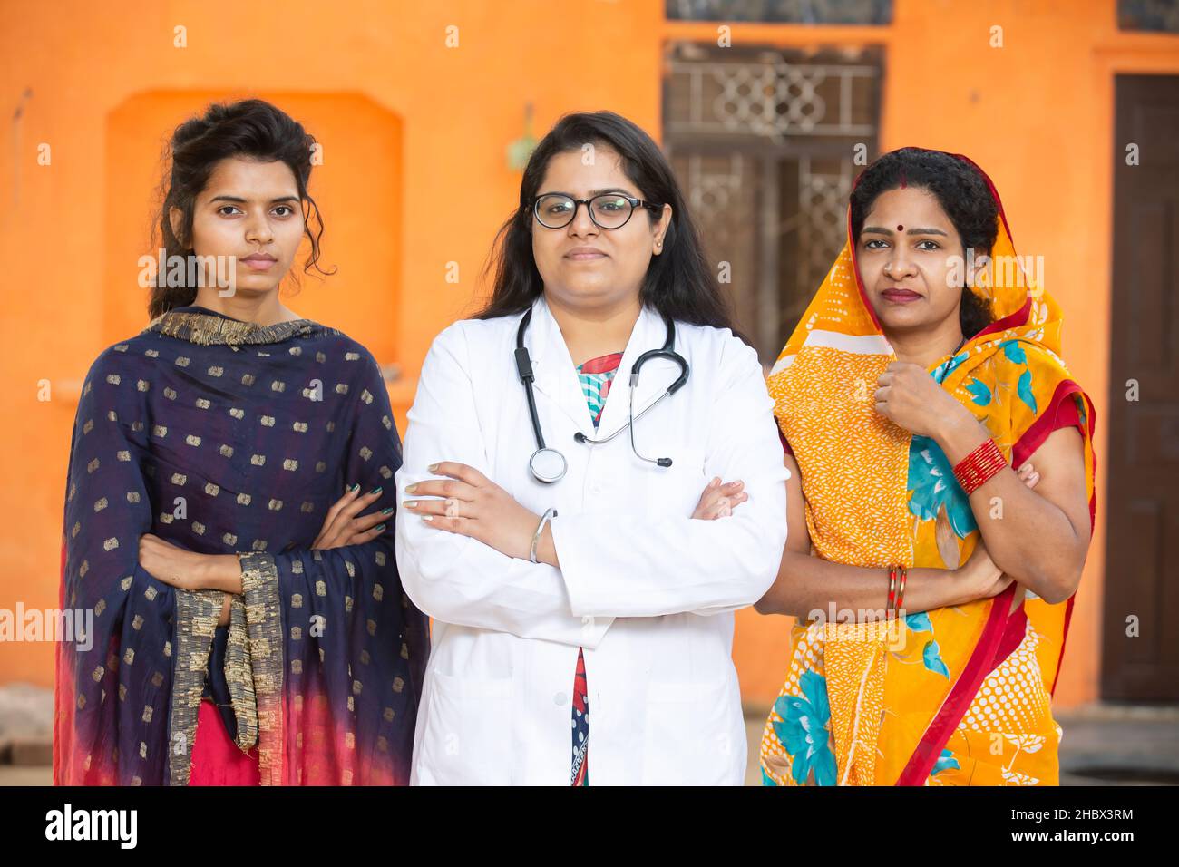 Ritratto di donna indiana in sari e sua figlia giovane in piedi con medico donna che indossa stetoscopio croce braccia, stanno guardando fotocamera, rurale Foto Stock