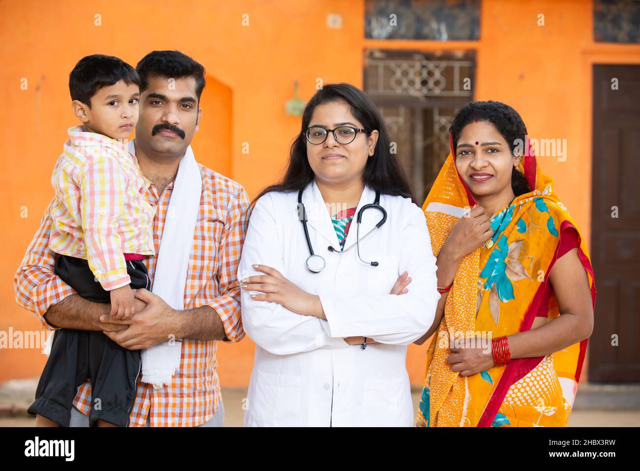 Ritratto della famiglia tradizionale indiana villaggio in piedi con medico femminile indossando stetoscopio croce braccia, genitore con il loro bambino, emozioni positive, Foto Stock